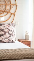 A neatly made bed with a patterned pillow, a woven wall hanging, and a bedside table with a decorative item.