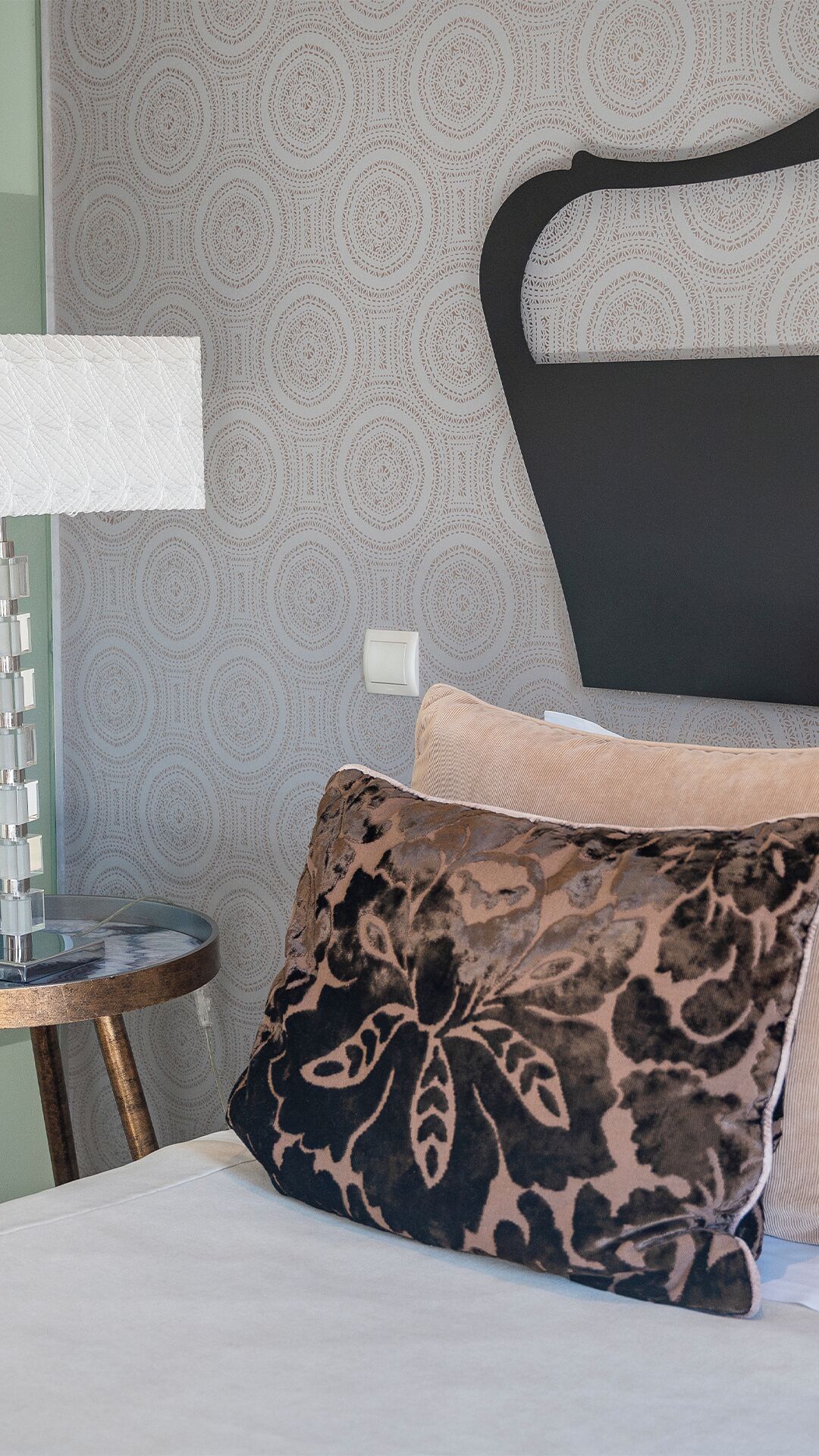 Cama bem arrumada com almofada estampada, mesa de cabeceira com abajur e parede com padrão circular.
