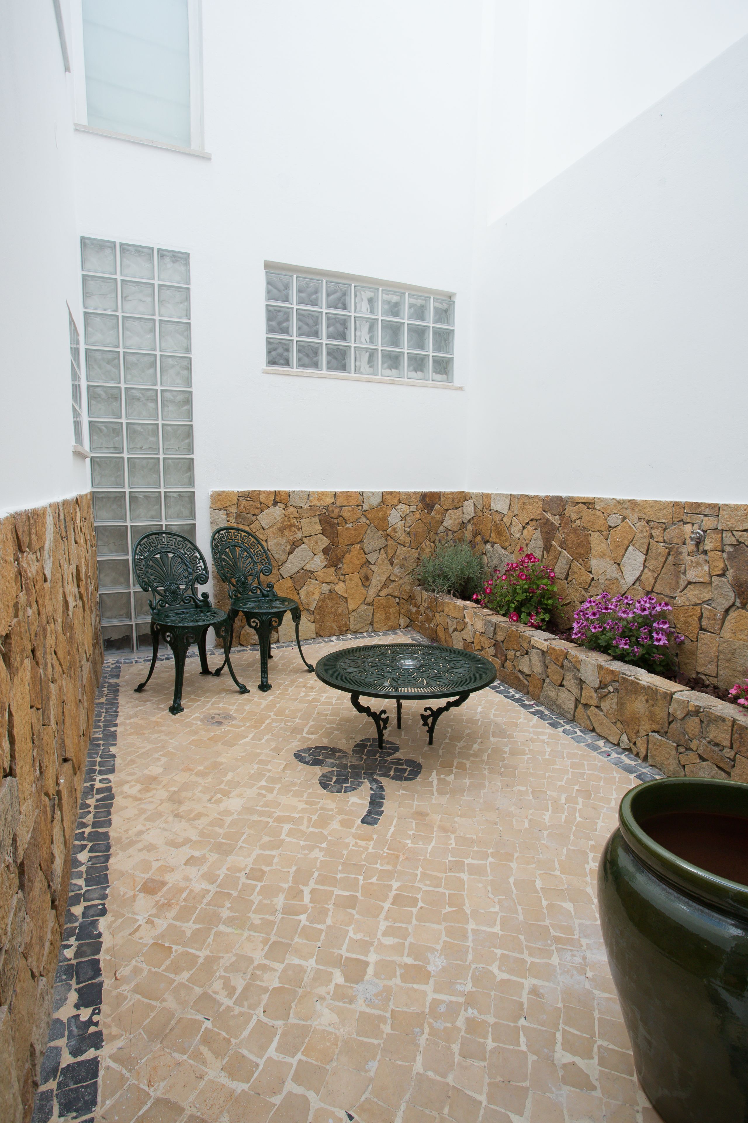 A courtyard with a stone wall, two metal chairs, a table, and a potted plant.
