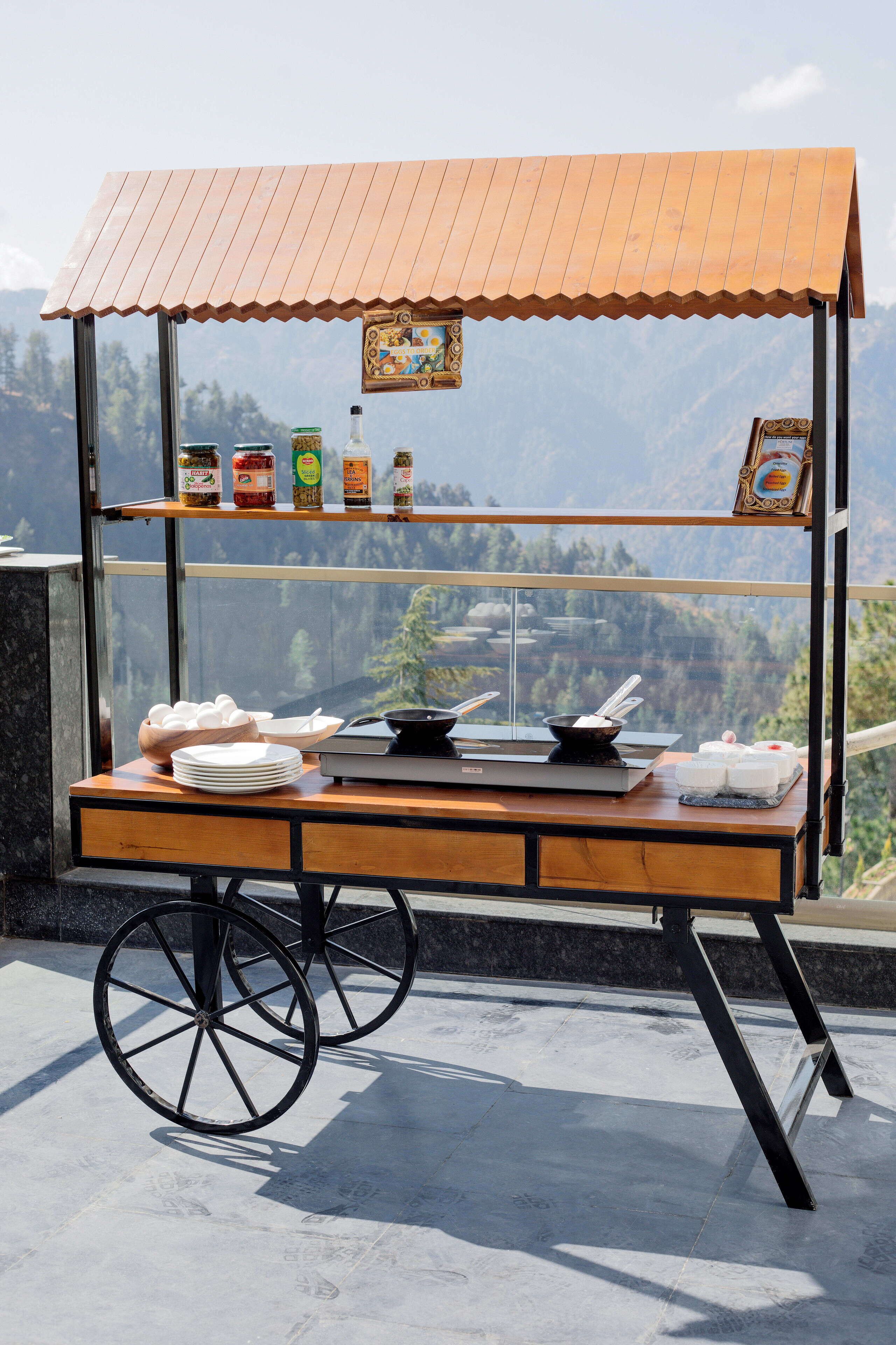 Un carrito de comida con cubierta de madera y estructura metálica, equipado con estantes para condimentos y un área de exhibición para alimentos.