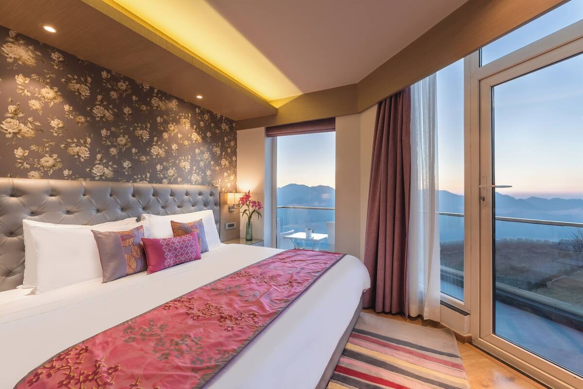Quarto de hotel com uma cama grande, vista para as montanhas e varanda com mesa e cadeiras.