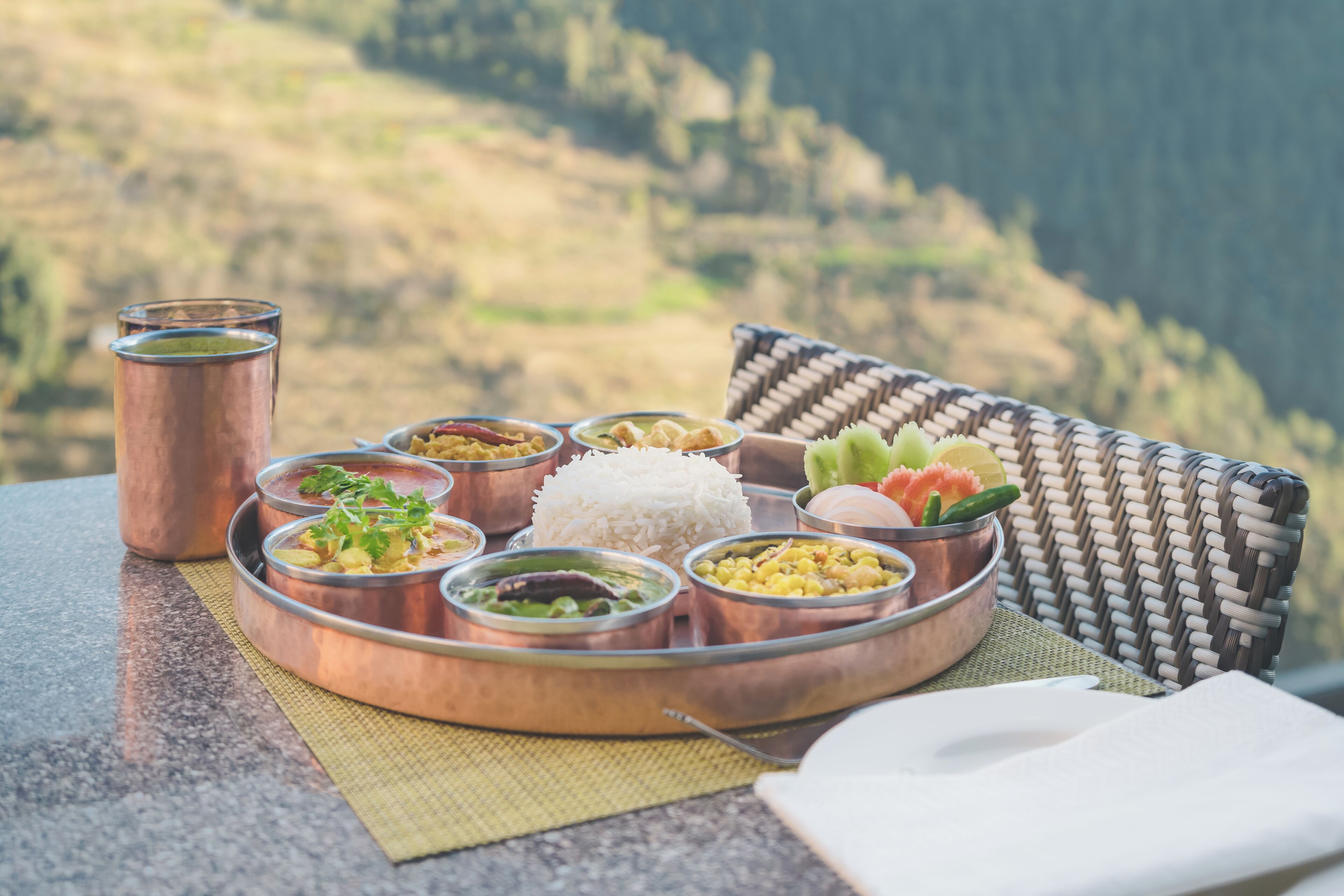 Un plateau dressé avec différents plats, une tasse et un panier tressé agrémenté d’un coussin, le tout présenté sur un fond pittoresque en plein air.