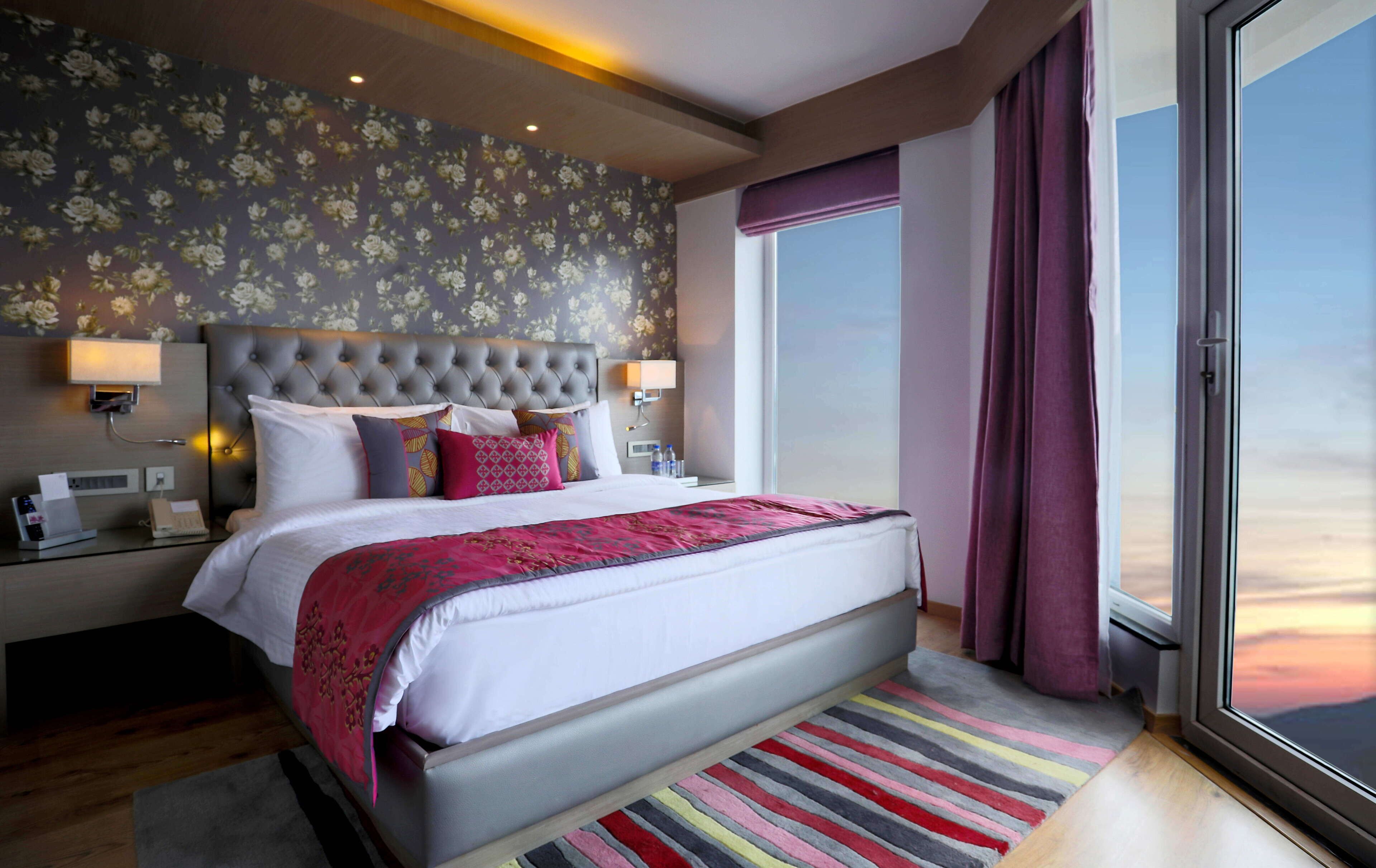 A modern hotel room with a large bed, a nightstand, a lamp, and a view of the sky through a large window.