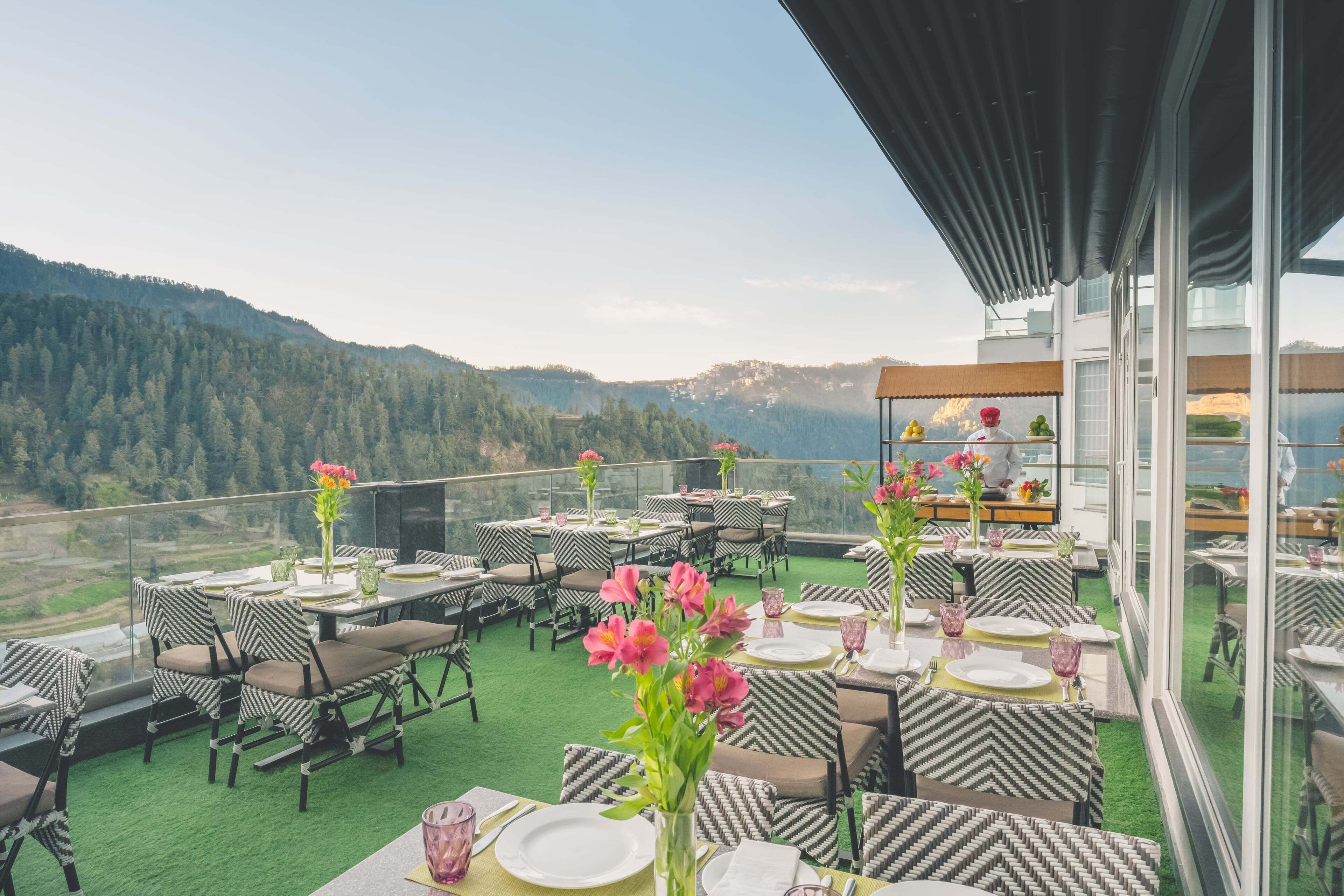 Un espace de restauration aménagé sur le toit, avec des tables et des chaises, offrant une vue sur les collines boisées, et une structure vitrée où un chef prépare les plats.