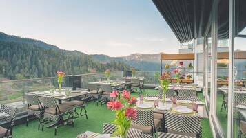 Un espace de restauration aménagé sur le toit, avec des tables et des chaises, offrant une vue sur les collines boisées, et une structure vitrée où un chef prépare les plats.