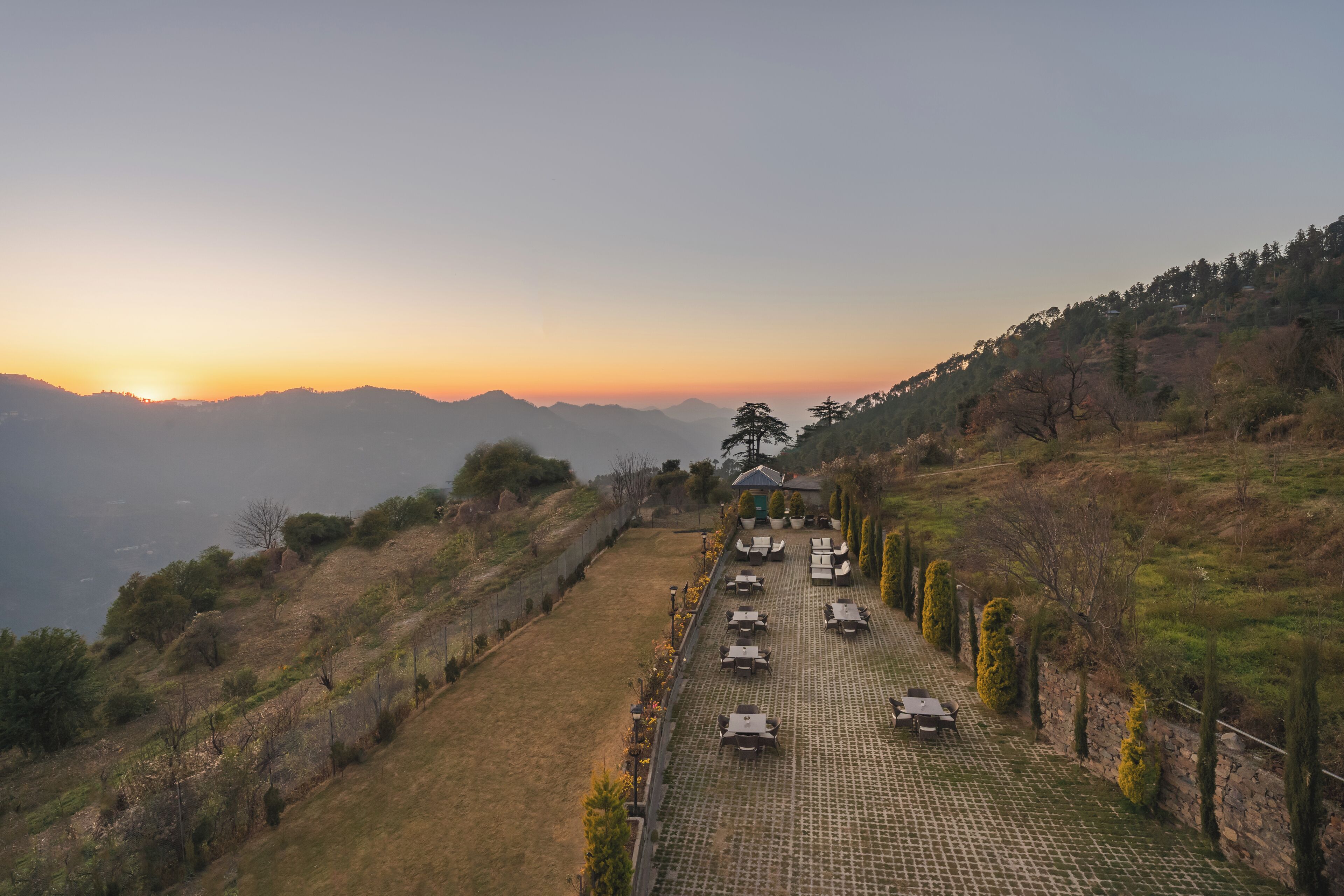A paved terrace with outdoor seating, overlooking a hilly landscape with a sunset in the background.