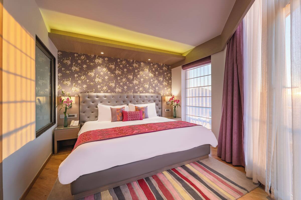 Um quarto espaçoso com uma cama grande, cabeceira estofada e vista para a cidade através de amplas janelas.