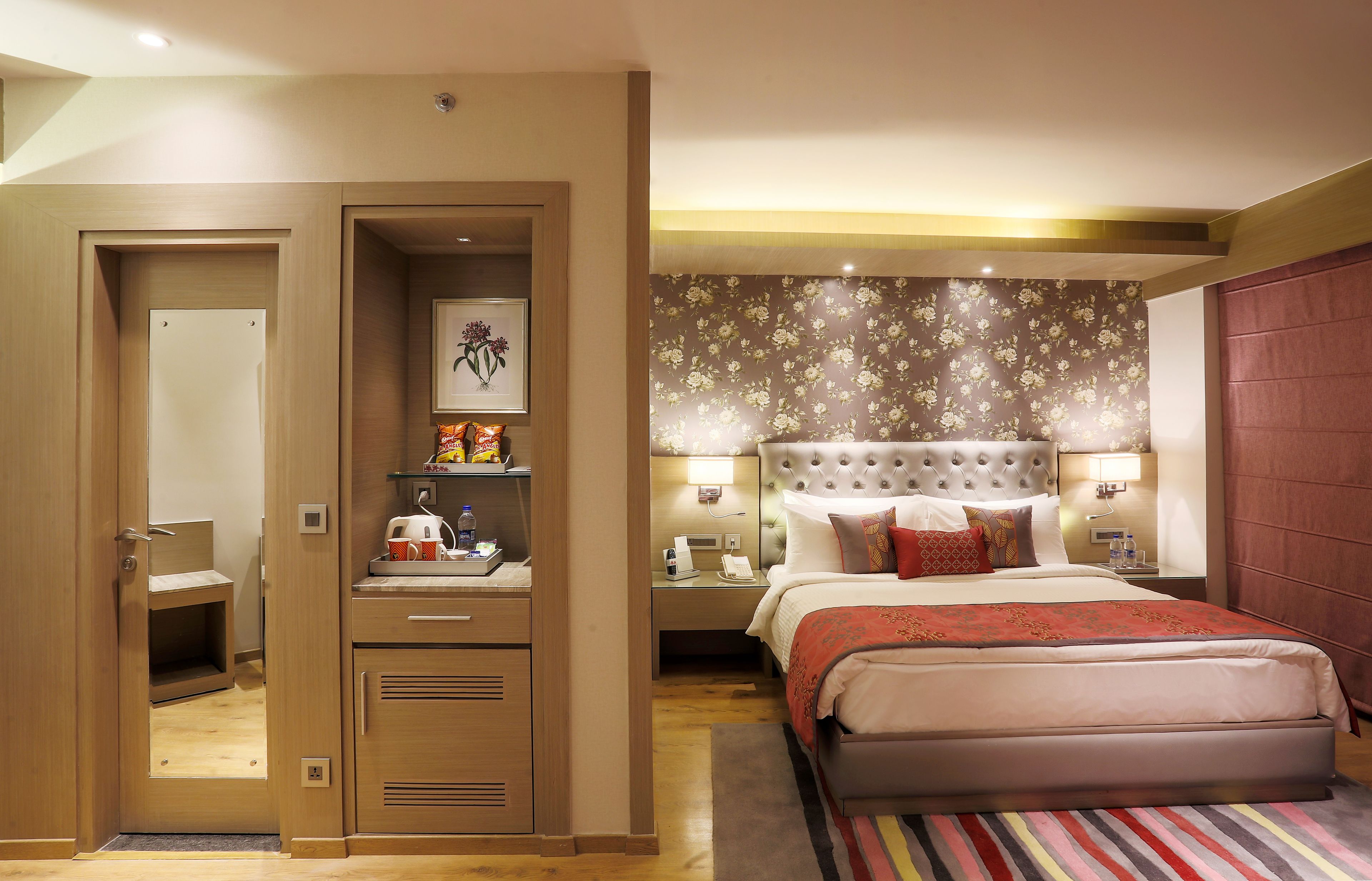 A hotel room with a bed, bedside tables, a nightstand, a wardrobe, a desk with a chair, and a door leading to another room.