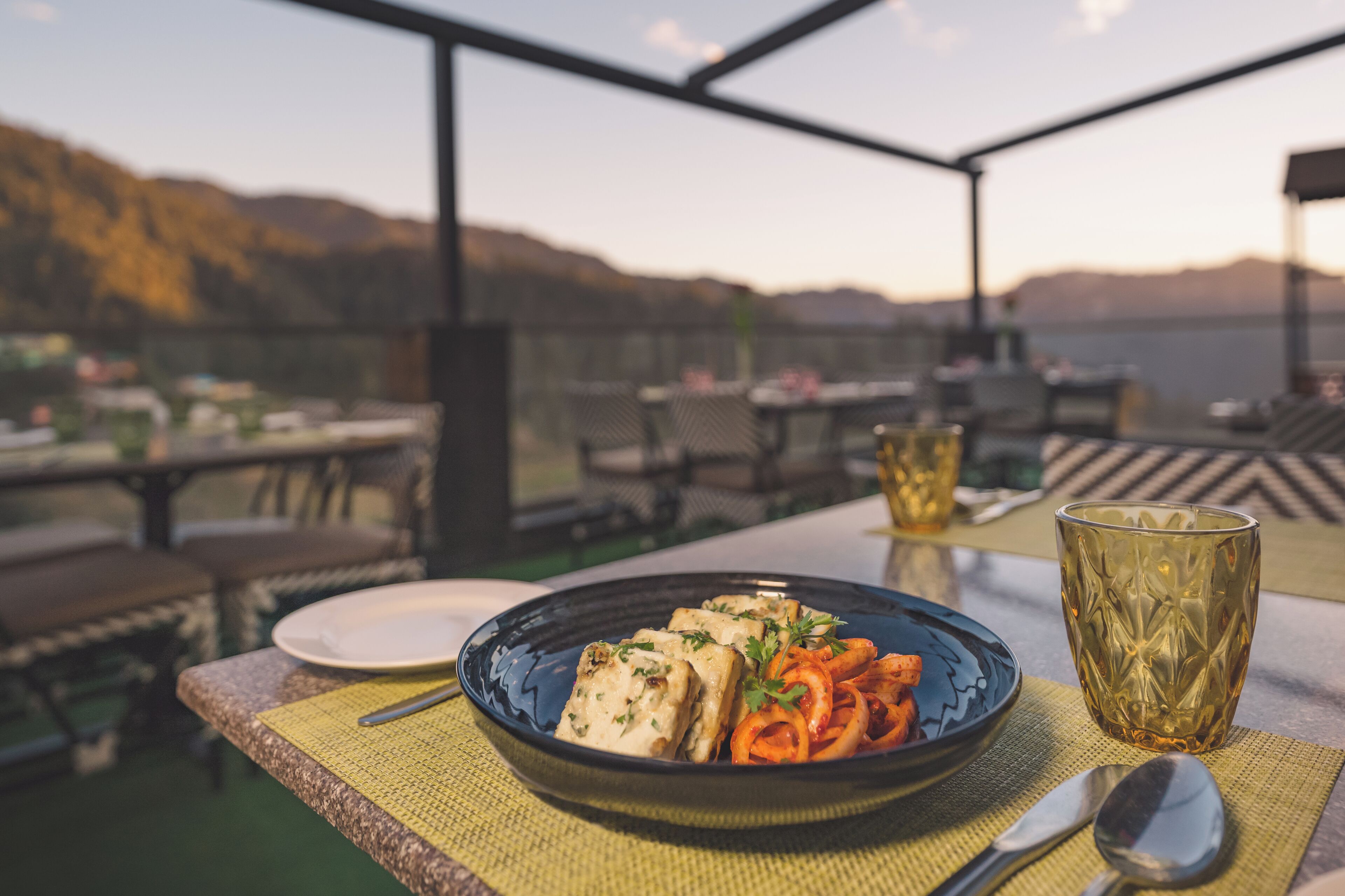 Een eettafel met een bord eten, een glas en bestek op een terras met uitzicht op bergen.