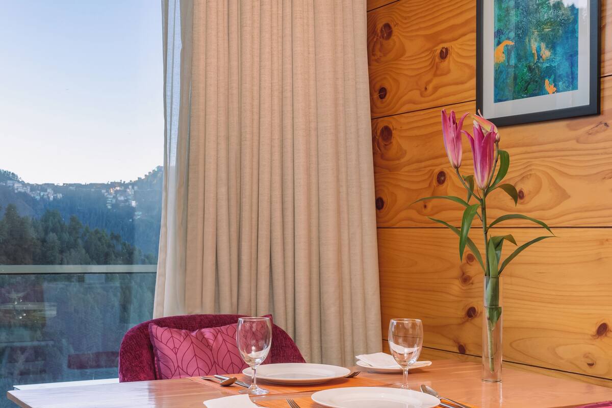 Mesa de jantar posta com pratos, copos e talheres, um vaso com uma flor, e vista para uma paisagem montanhosa através de uma janela.
