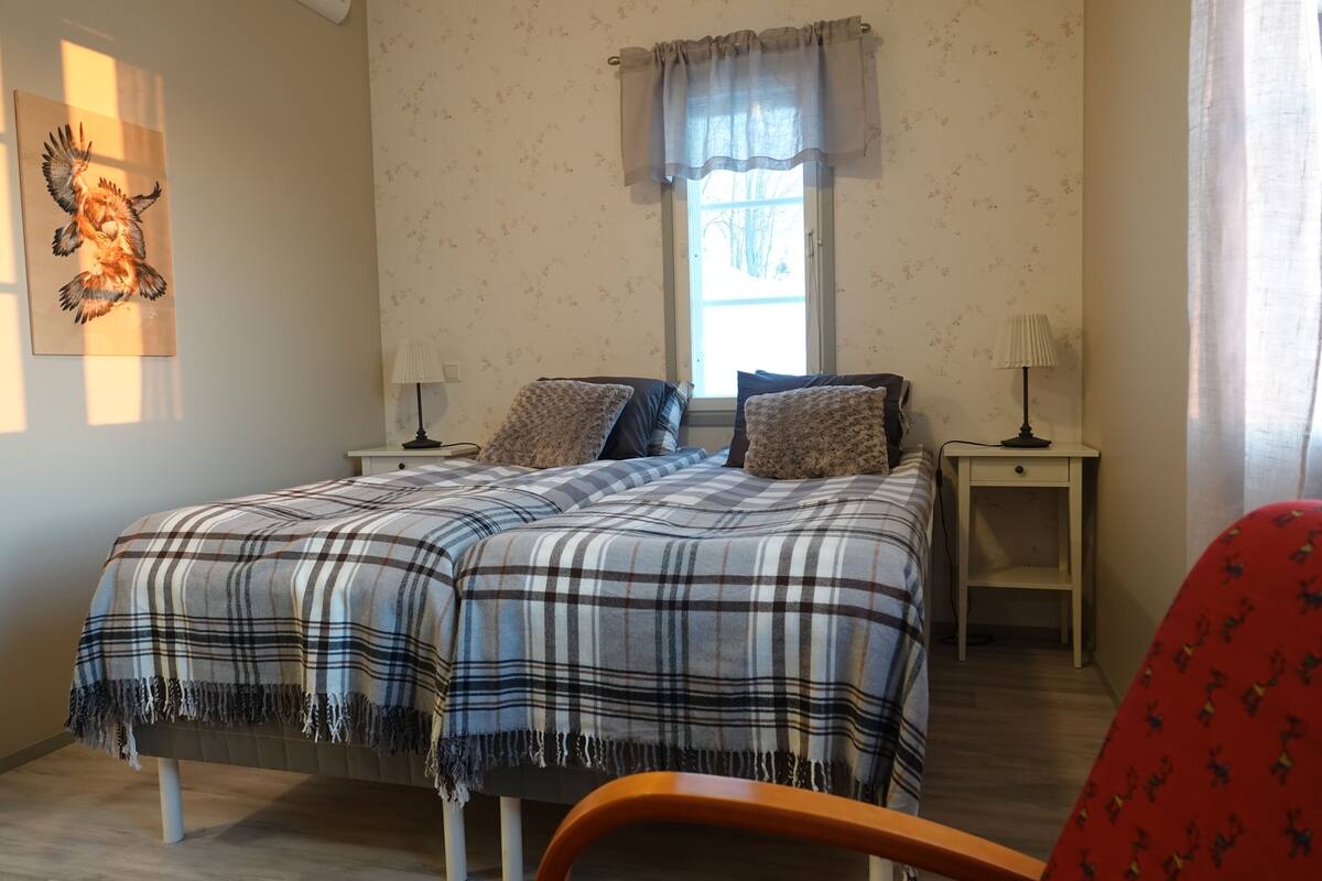 A bedroom with a double bed, two bedside tables, a window with curtains, a wall-mounted artwork, and a red chair.
