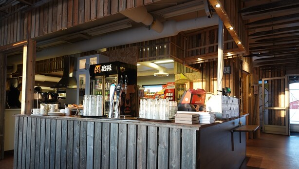 Un comptoir en bois rustique, avec une machine à café, des tasses et une enseigne sur laquelle on peut lire : « TAVATTA GLUHTA ».
