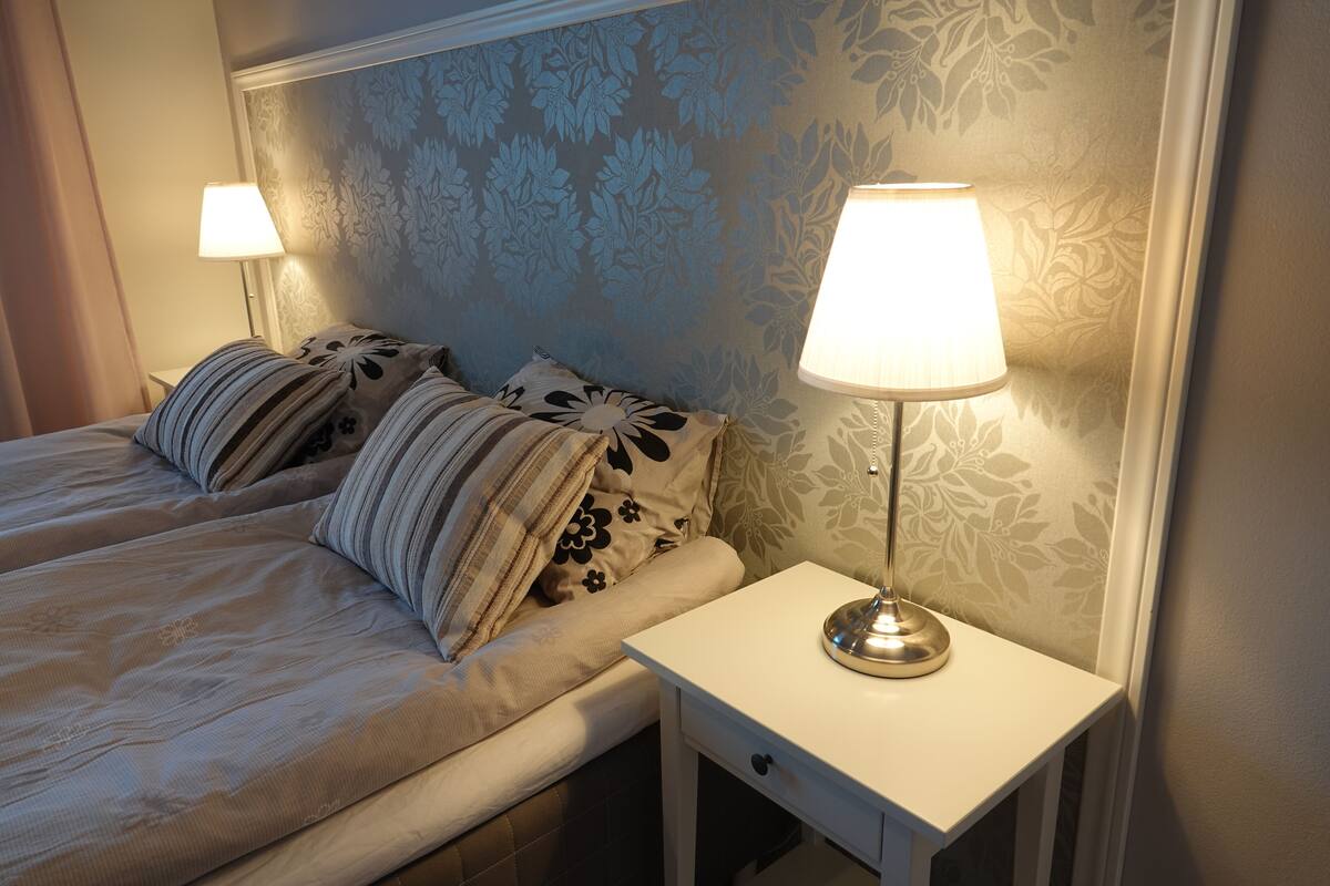 A neatly made bed with striped pillows, a bedside lamp, and a floral-patterned wall.