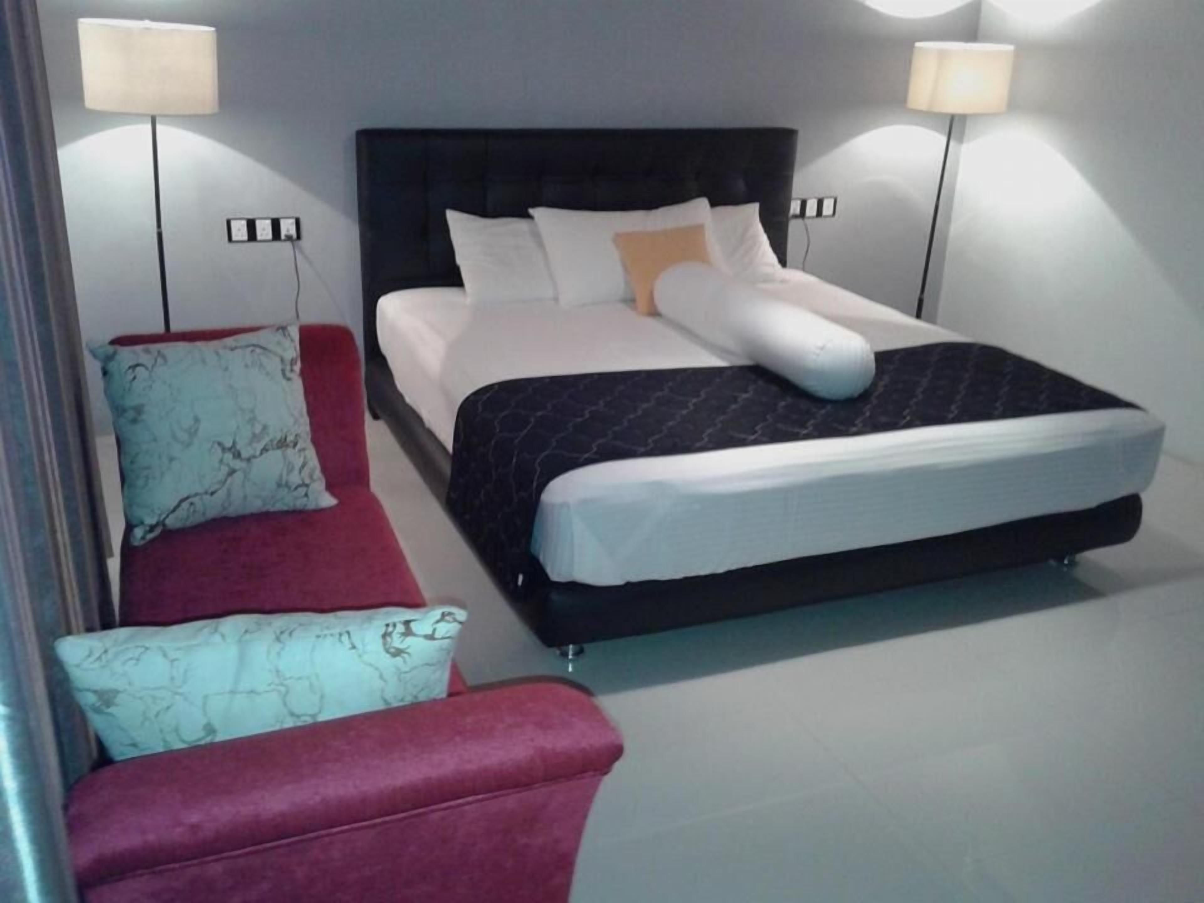 A modern hotel room with a large bed, two bedside lamps, a red sofa, and a patterned armchair.