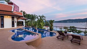 Ein Pool mit geschwungener Form, umgeben von Liegestühlen und Topfpflanzen, mit Blick auf ein Gewässer, in dessen Ferne Land und Boote liegen.