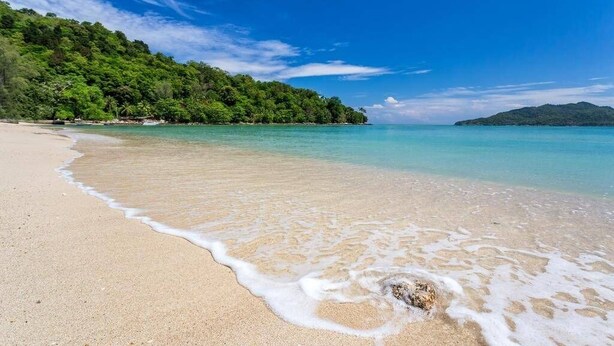 Ein Strand mit klarem türkisblauem Wasser, sandigem Ufer und einem bewaldeten Hügel im Hintergrund.