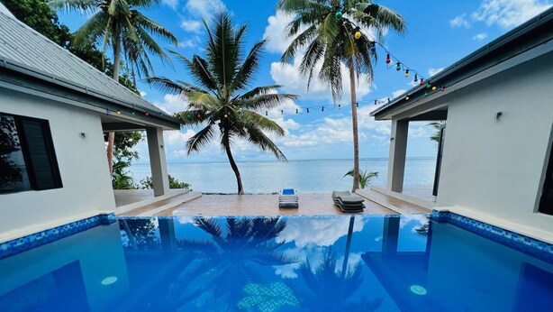 A pool with a clear view of the ocean, surrounded by palm trees and a white building.