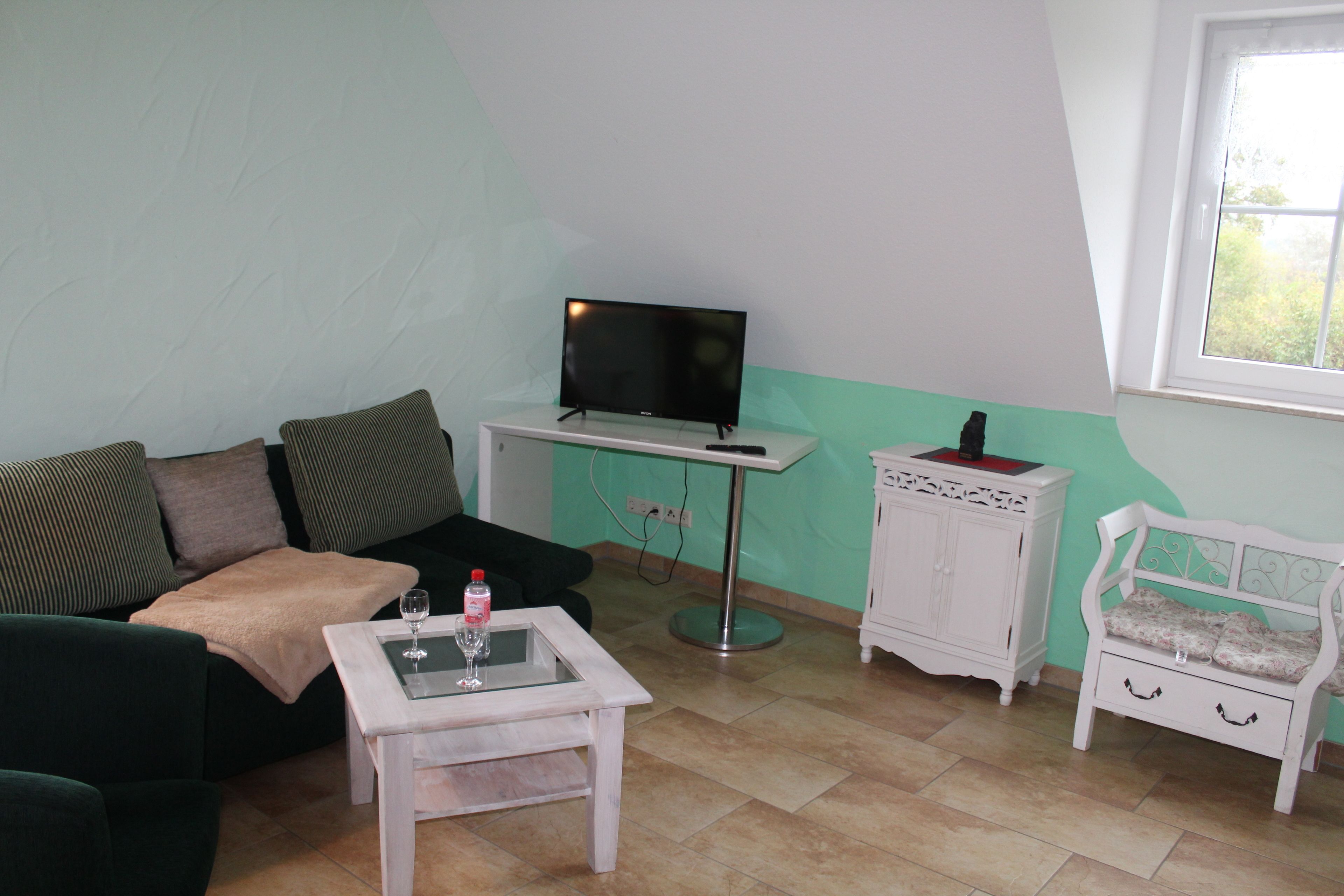 Un salon avec un canapé vert, une table basse blanche avec un plateau en verre, un téléviseur posé sur un meuble, une armoire blanche et une fenêtre donnant sur un paysage verdoyant.