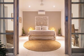 Relax in this serene space with a central bed, bedside tables, and a large rug, surrounded by lush greenery and a beautiful wall hanging.
