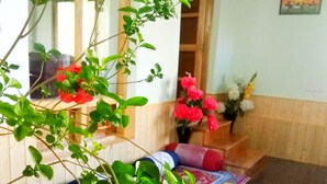 Ein Zimmer mit einem langen Holztisch, bunten Teppichen mit Muster und einem Blumentopf mit grünen Blättern und roten Blüten.