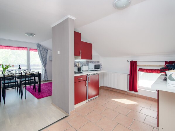 Cocina compacta con armarios rojos, microondas y refrigerador pequeño. Cuenta con un comedor con mesa y sillas, además de un ventanal con cortinas.