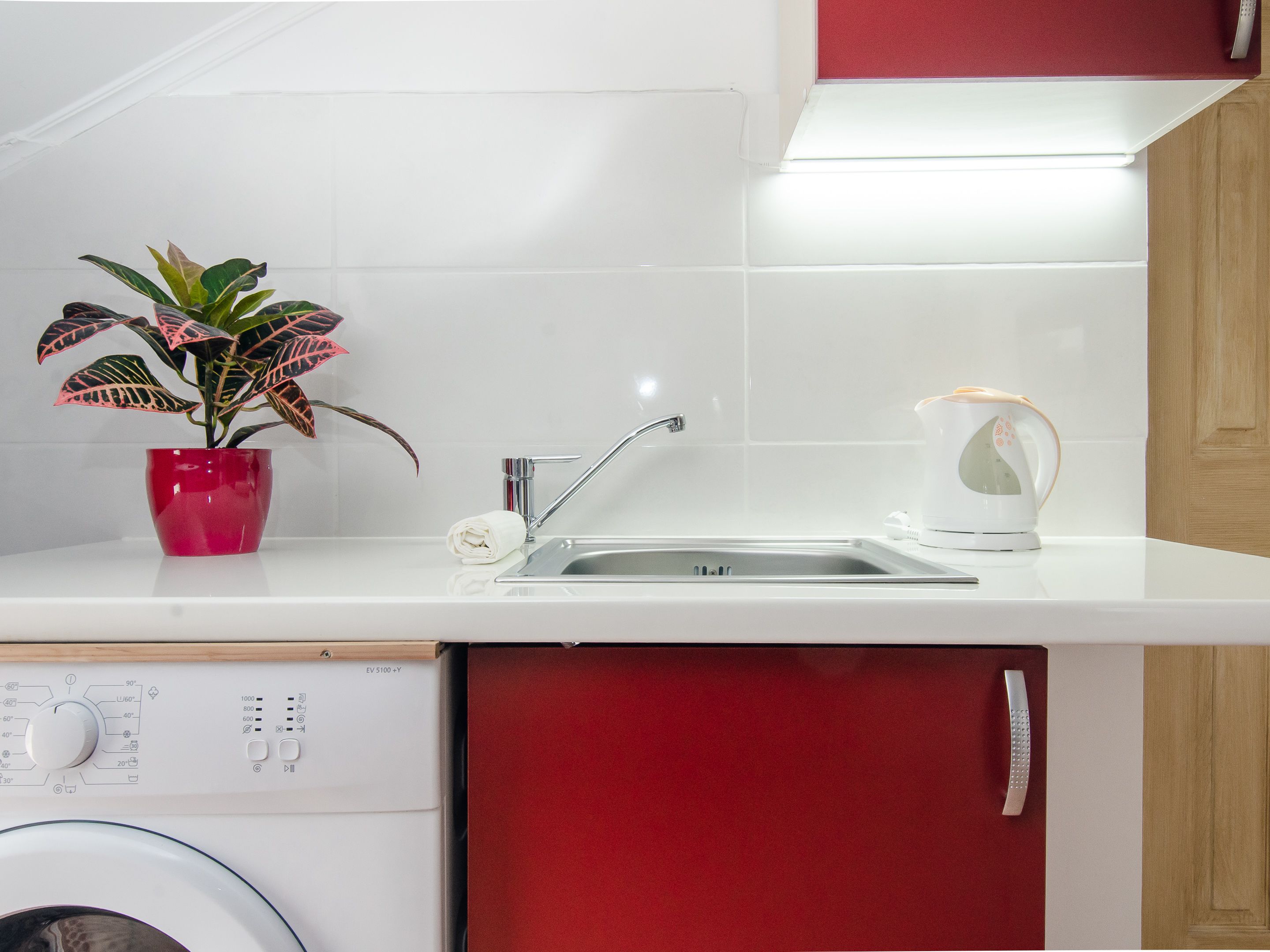 A kitchen with a white countertop, a stainless steel sink, a red cabinet, a white appliance with a control panel, and a potted plant.