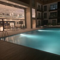 An indoor swimming pool with clear water, surrounded by a tiled deck and a glass wall with a restaurant.
