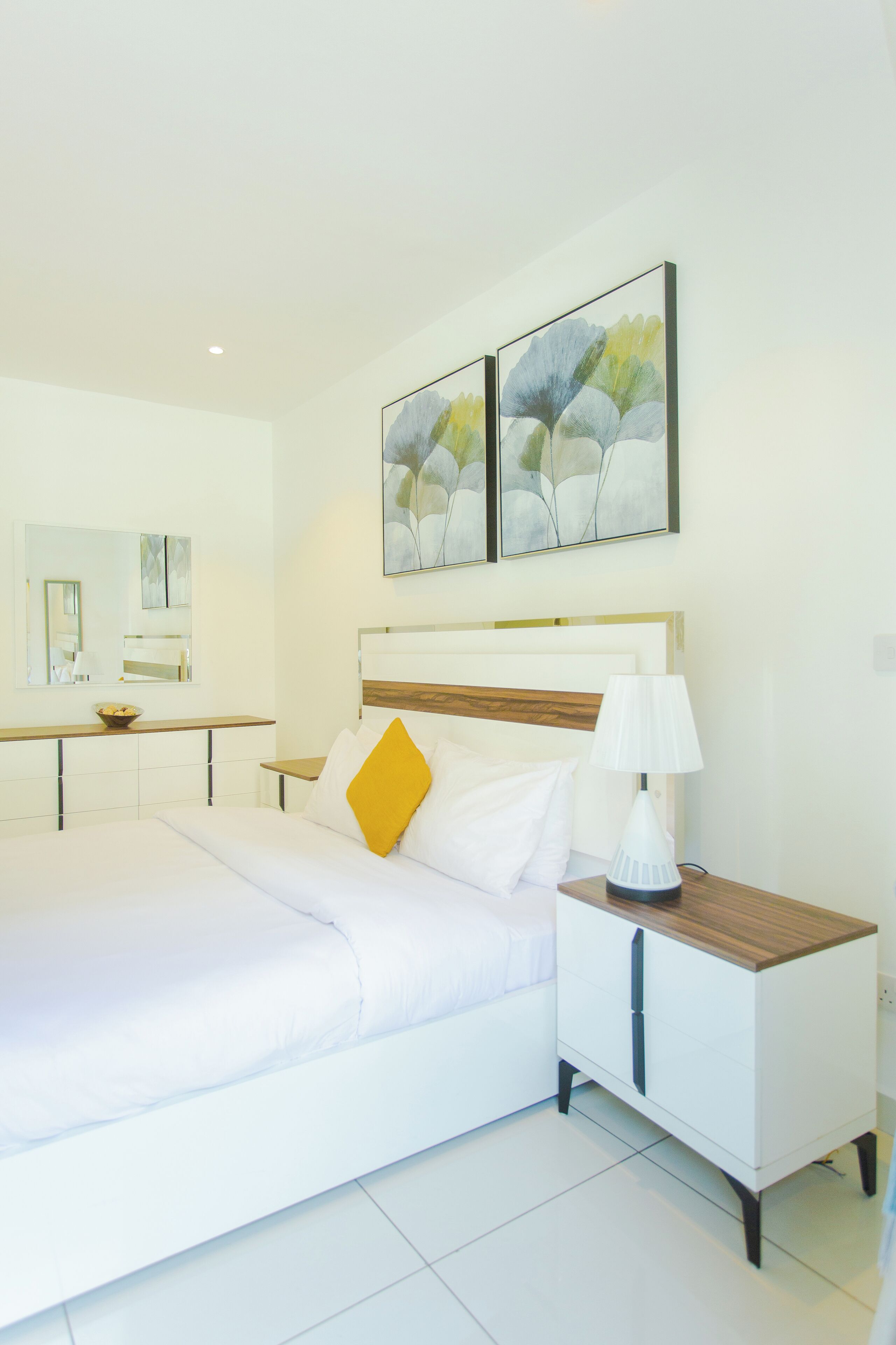 A modern hotel room with a large bed, a bedside table with a lamp, a small wooden nightstand, and a mirror above the bed.