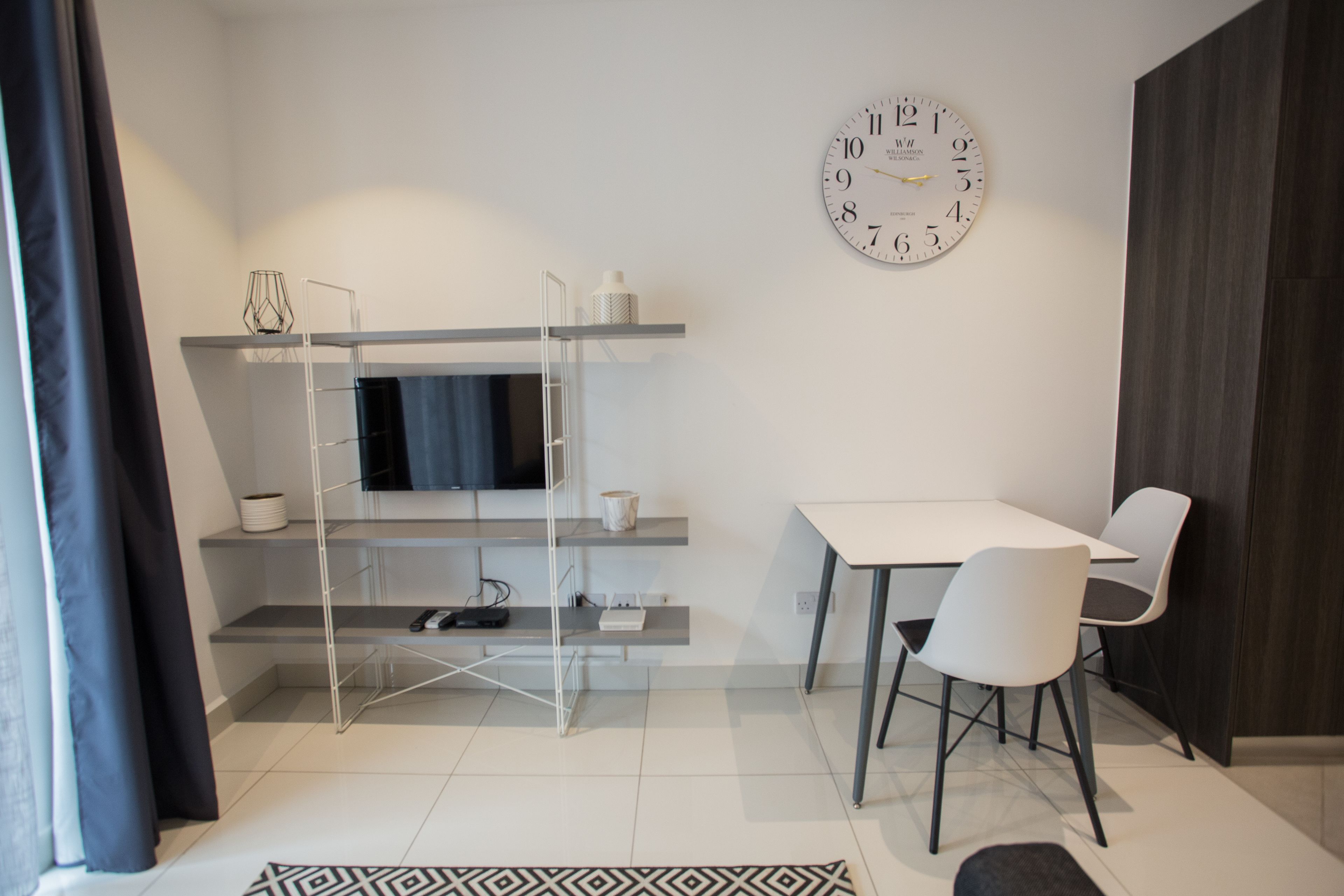 Een moderne kamer met een televisie op een plank, een eettafel met stoelen en een grote wandklok.