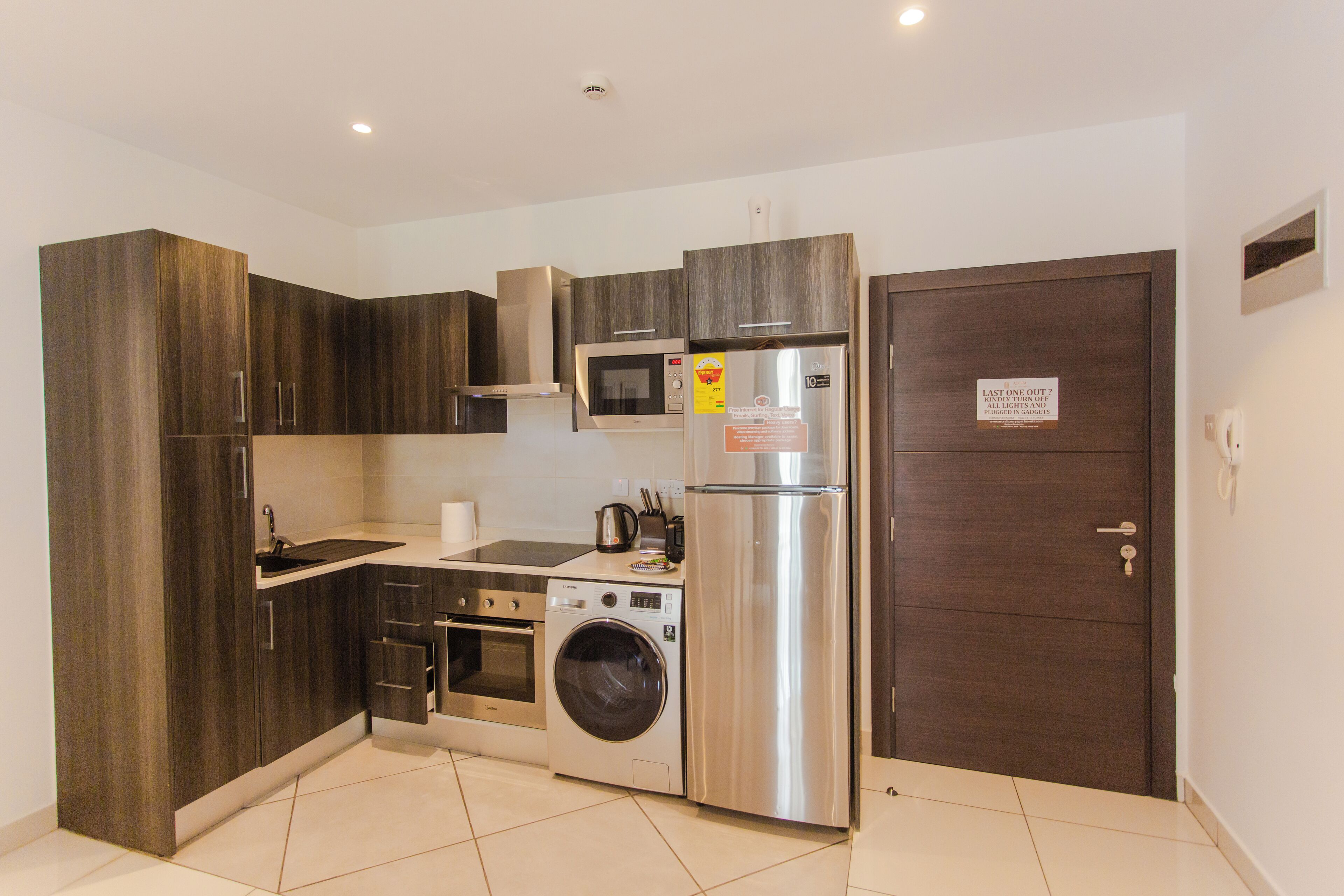 A compact kitchen with dark wood cabinets, a stainless steel refrigerator, a microwave, an oven, and a washing machine.