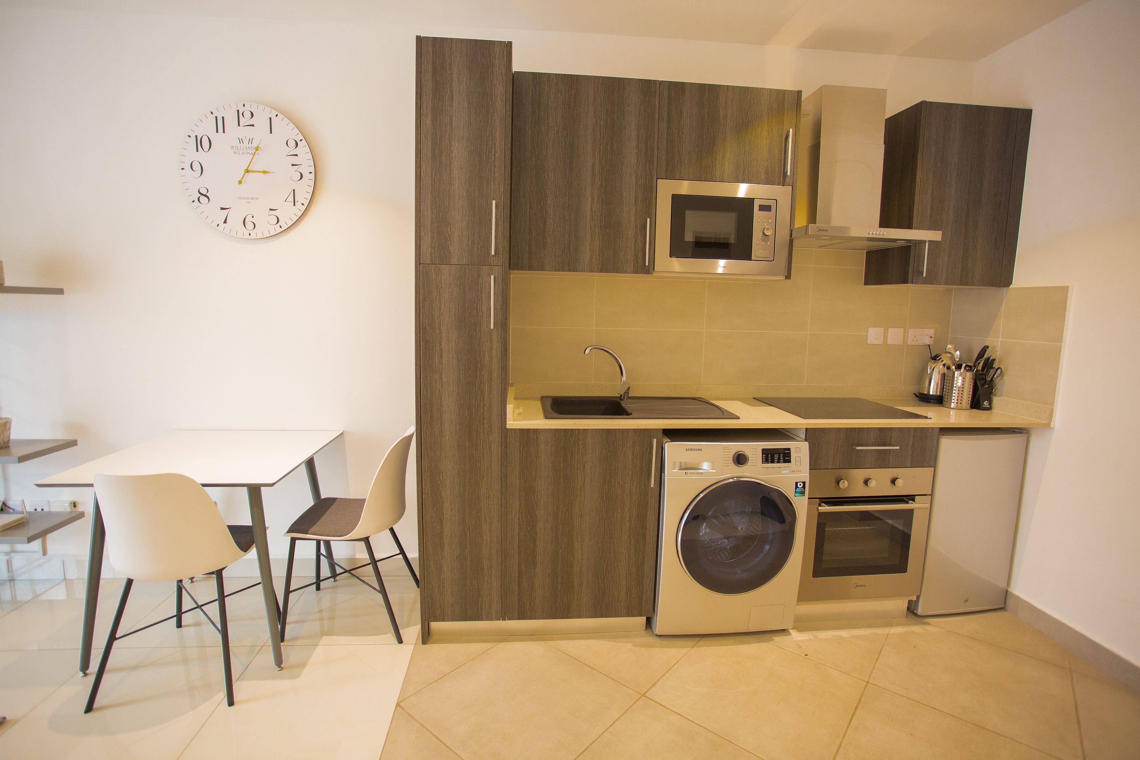 A compact kitchen with a microwave, oven, and washing machine, a dining area with chairs, and a wall clock.