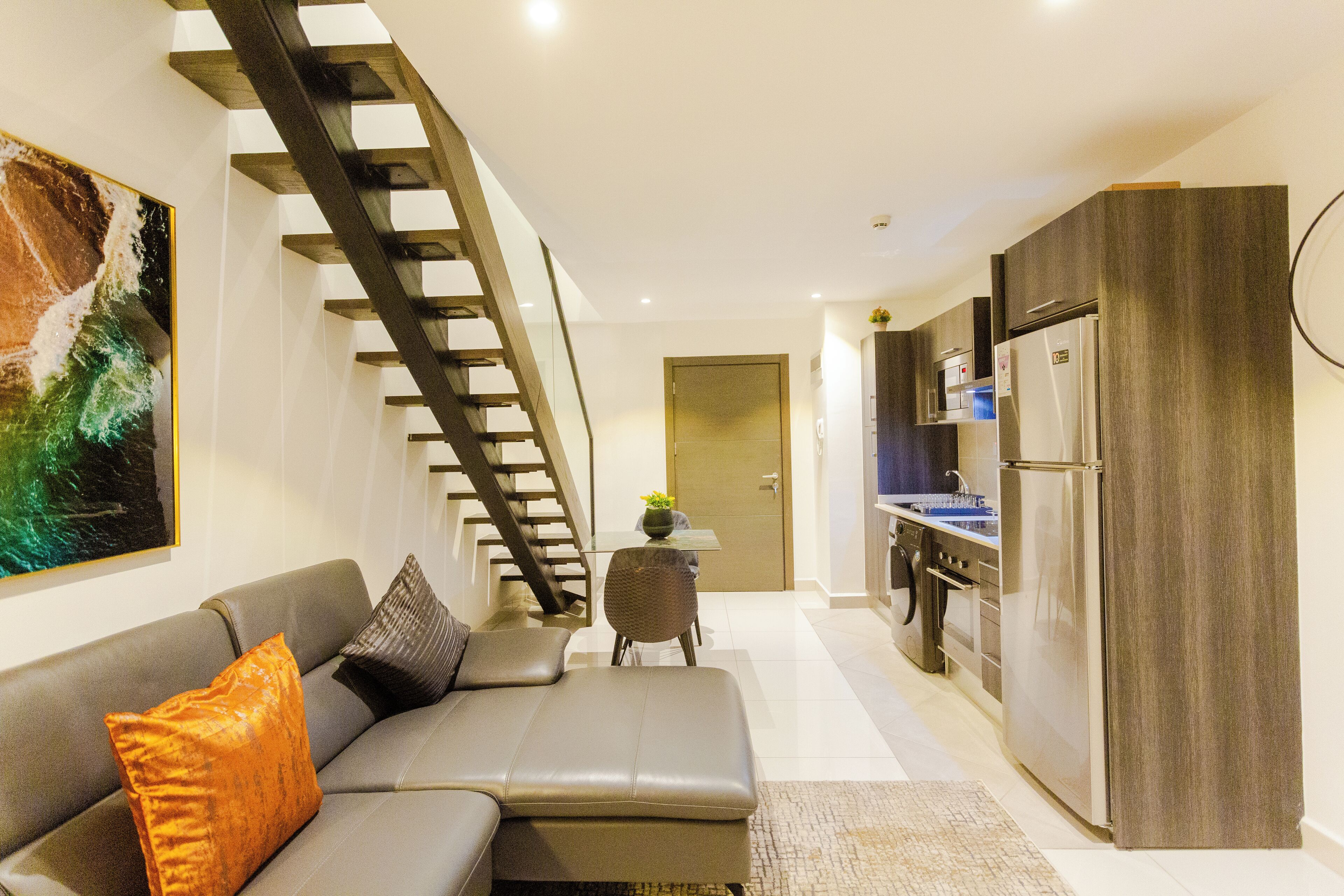 Ein modernes Wohnzimmer mit grauem Sofa, einer Treppe, einer Küche mit Edelstahlkühlschrank und einer Tür zu einem weiteren Raum.