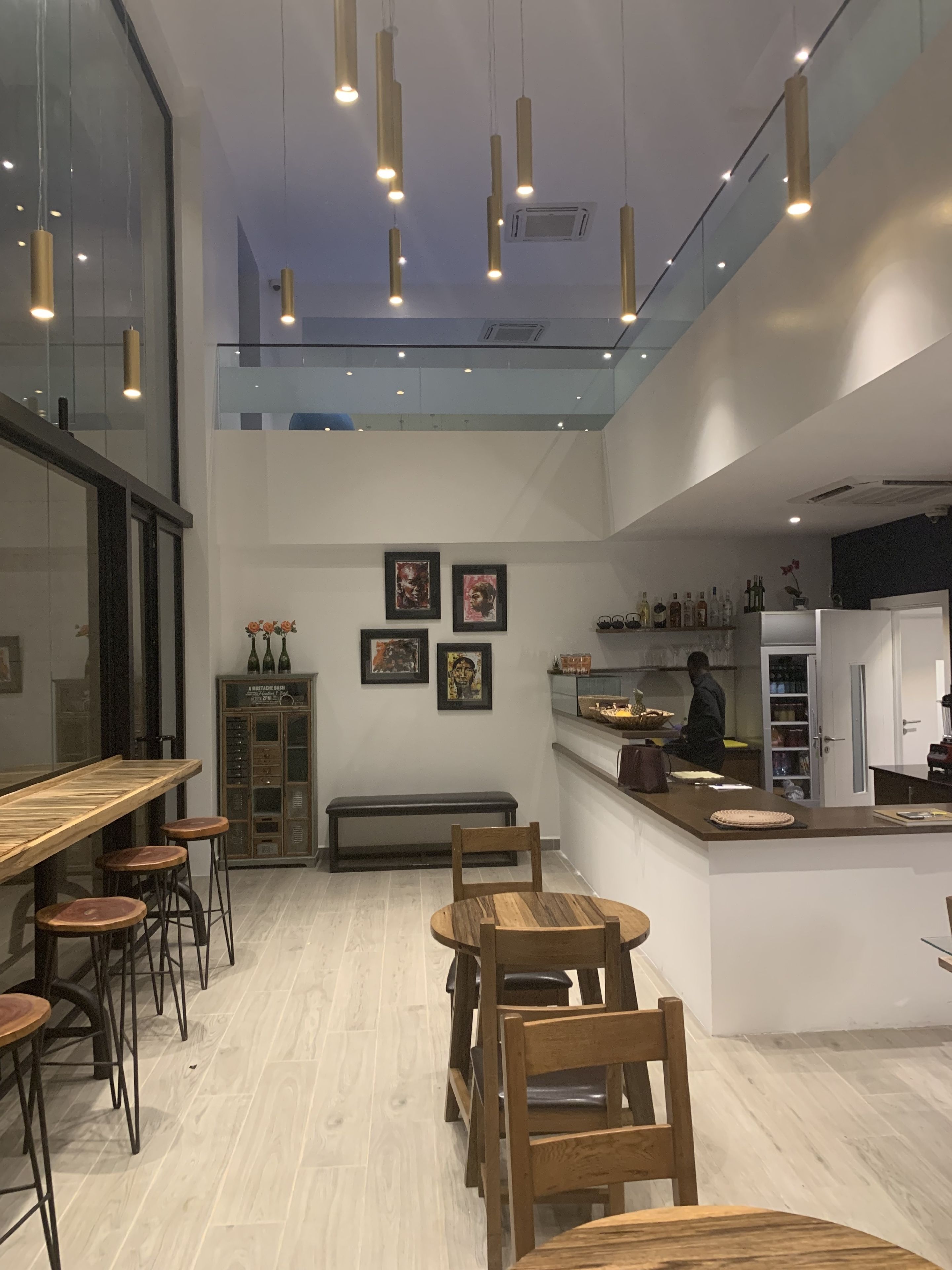 A modern restaurant interior with wooden tables and chairs, a high ceiling with pendant lights, and a person working at the counter.