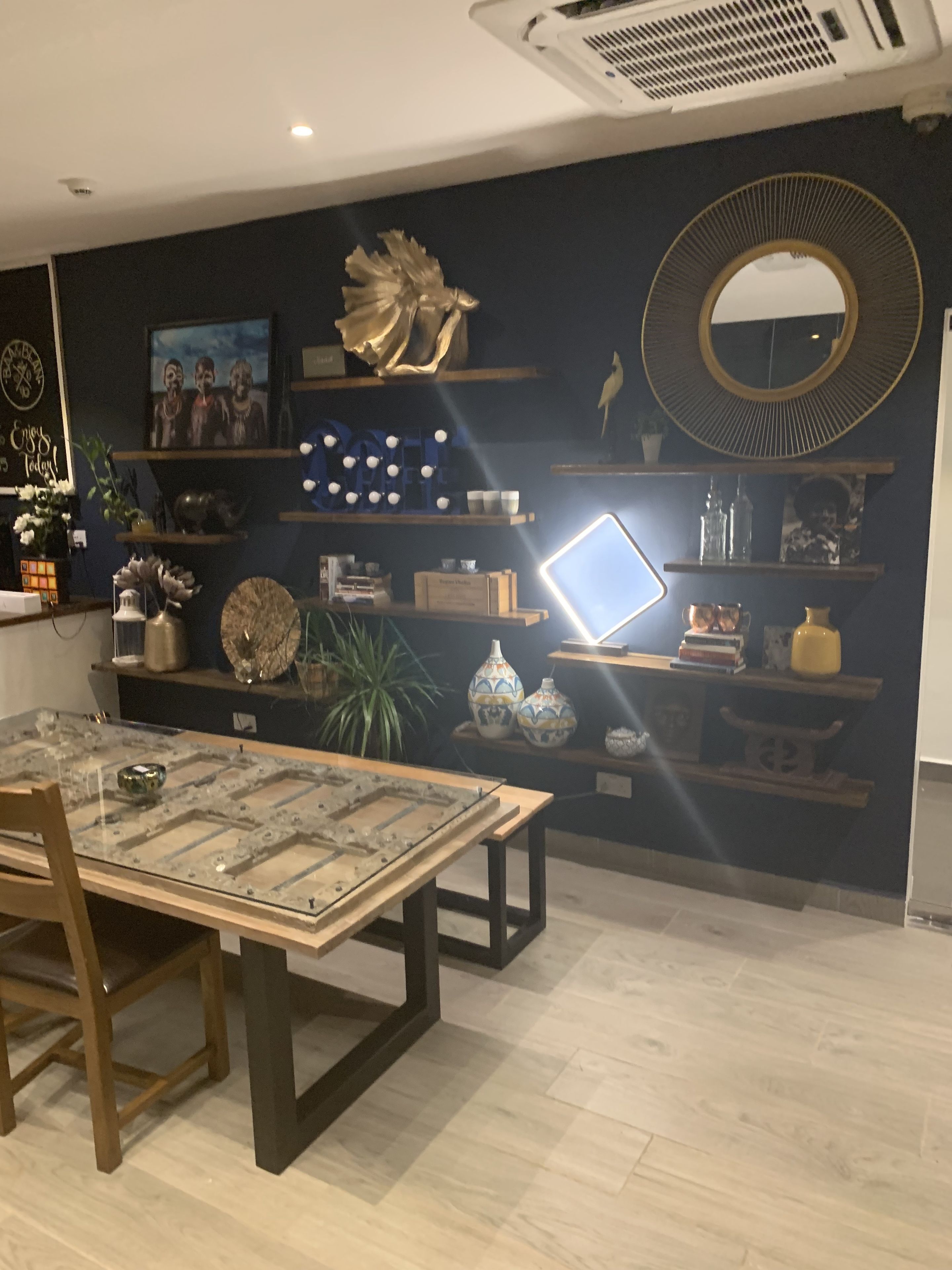 A dining area with a glass table, chairs, and a dark wall with shelves displaying decorative items and a laptop.