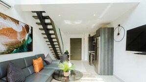 Een moderne woonkamer met een grijze bank, een glazen salontafel met een plant, een trap en een keukenhoek met een tv aan de muur.