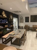 A modern dining area with a long table, wooden chairs, and a glass-top table. There are shelves with decorative items and a chalkboard menu on the wall.