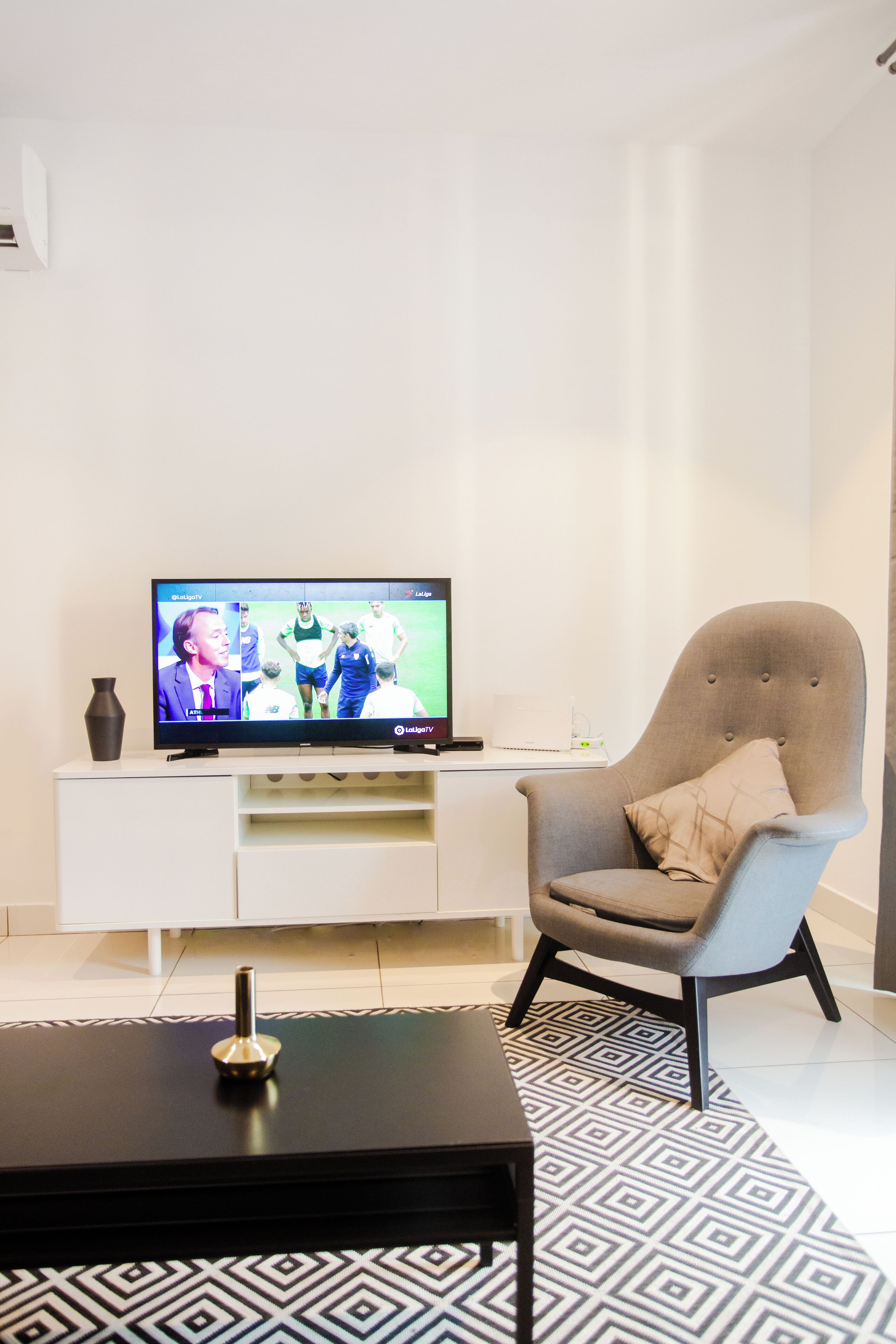 Una sala de estar moderna con un televisor de pantalla plana sobre un mueble blanco, un sillón gris con un cojín, una mesa de centro negra y una alfombra estampada.