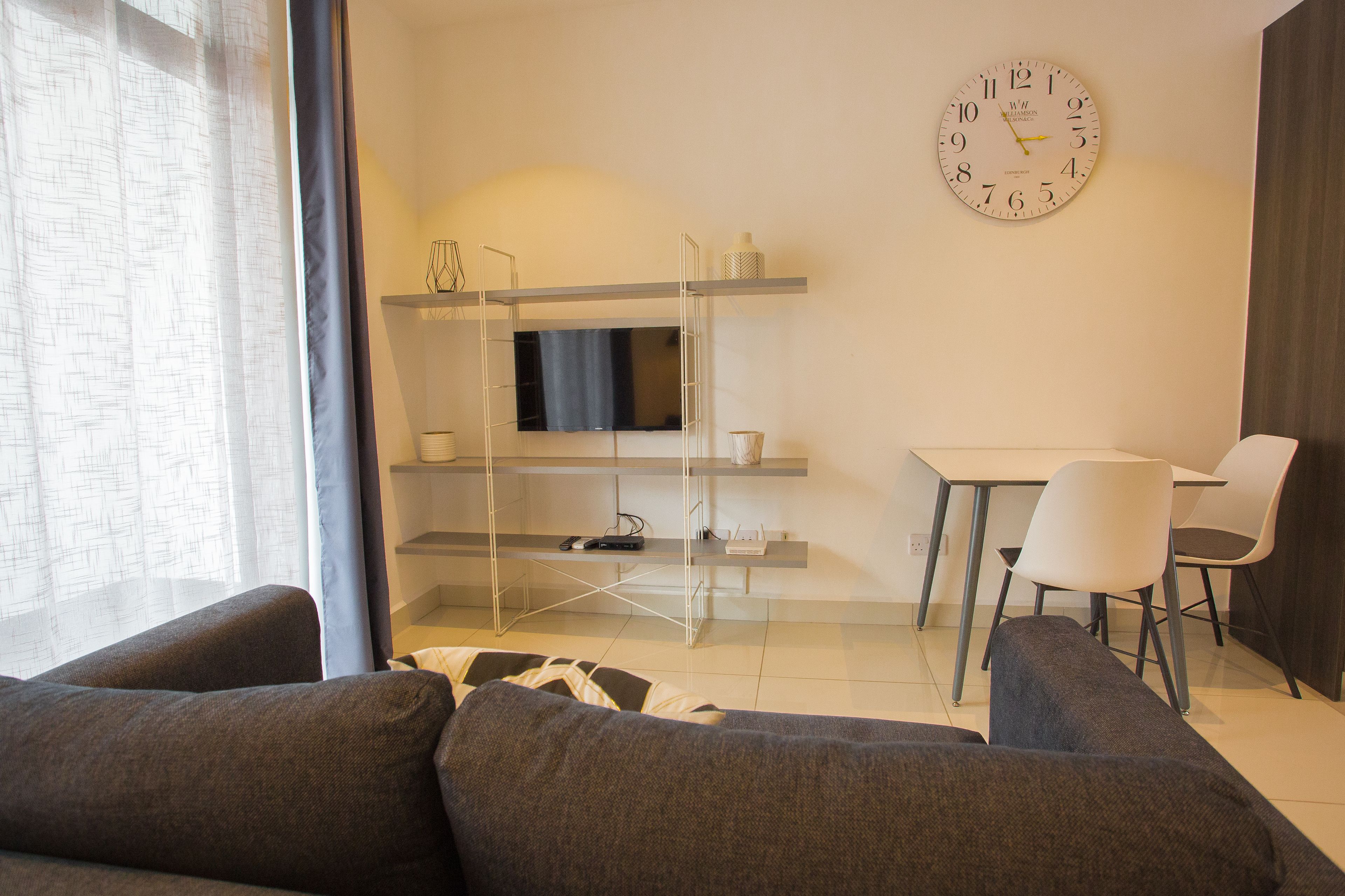 A modern living room with a sofa, a TV on a shelf, a dining table with chairs, and a clock on the wall.