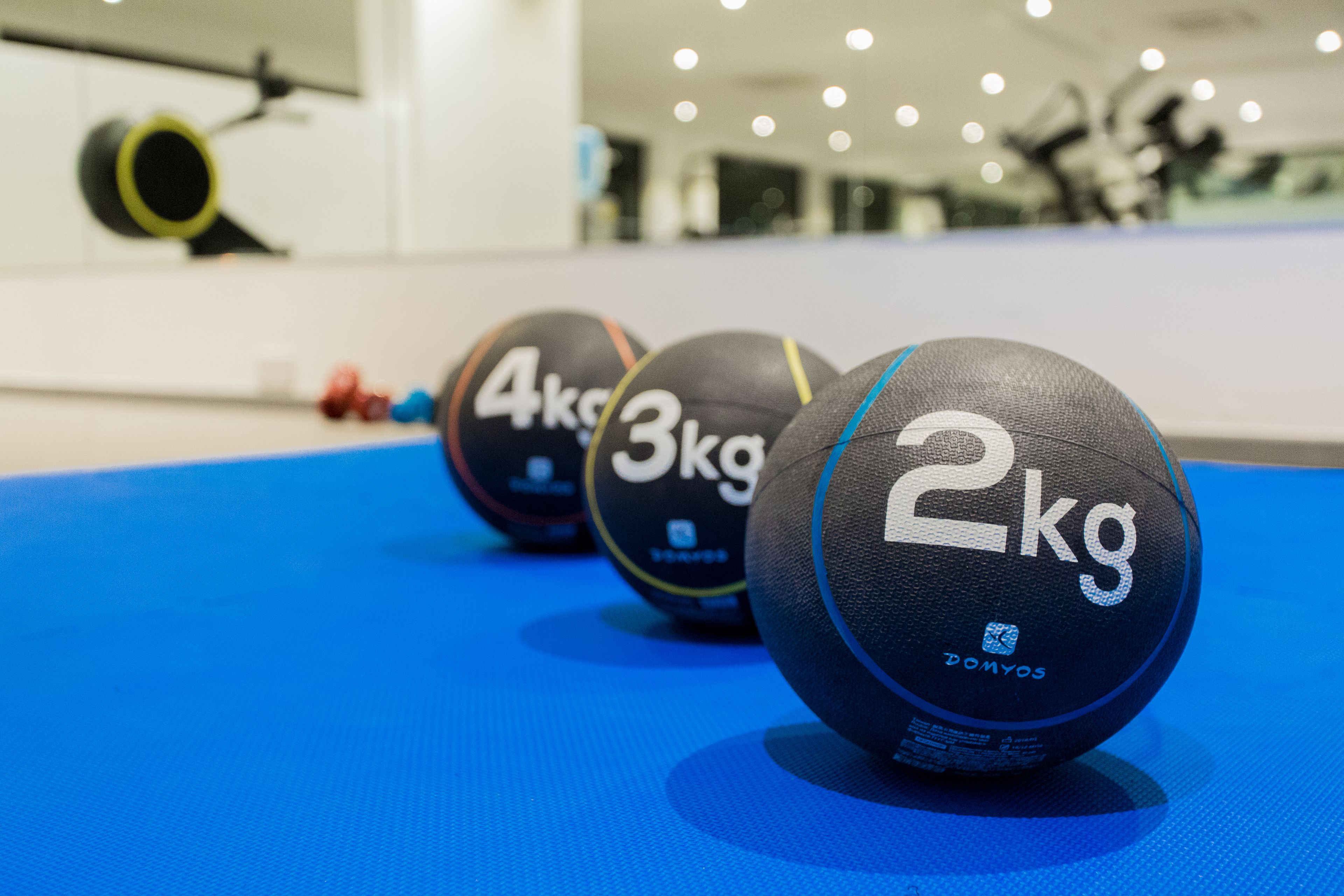 Tres pelotas de ejercicio marcadas con pesas sobre una colchoneta azul en un gimnasio.