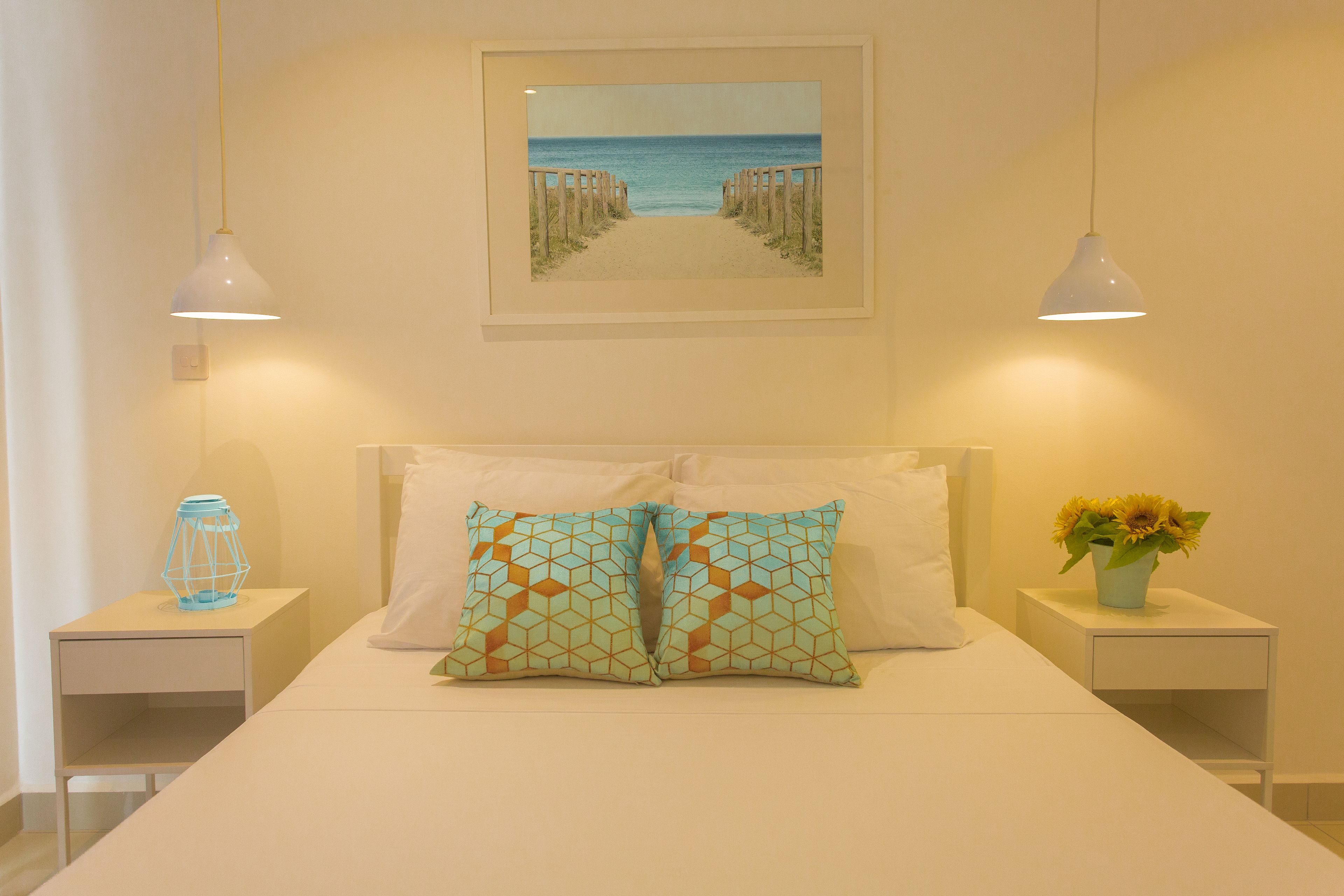 A bedroom with a bed, two bedside tables, a nightstand, a framed picture of a beach, and a vase with flowers.
