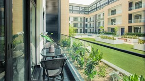 Ein Balkon mit Glastür, Tisch und Stühlen mit Blick auf einen Innenhof, der mit Pflanzen und einem Gebäude im Hintergrund gestaltet ist.