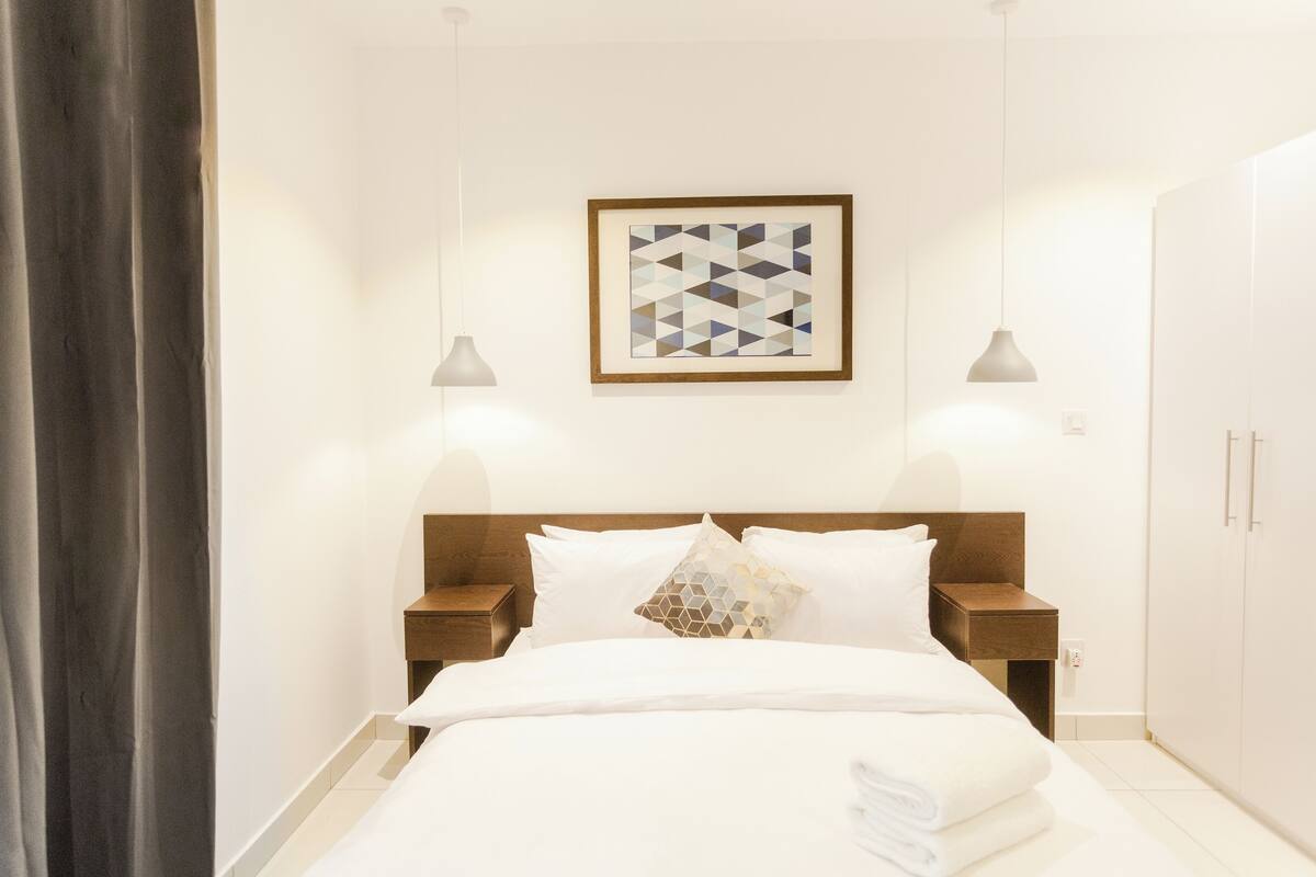 Une chambre d’hôtel comprenant un lit, deux tables de chevet, une petite table, une œuvre d’art géométrique encadrée et une armoire.