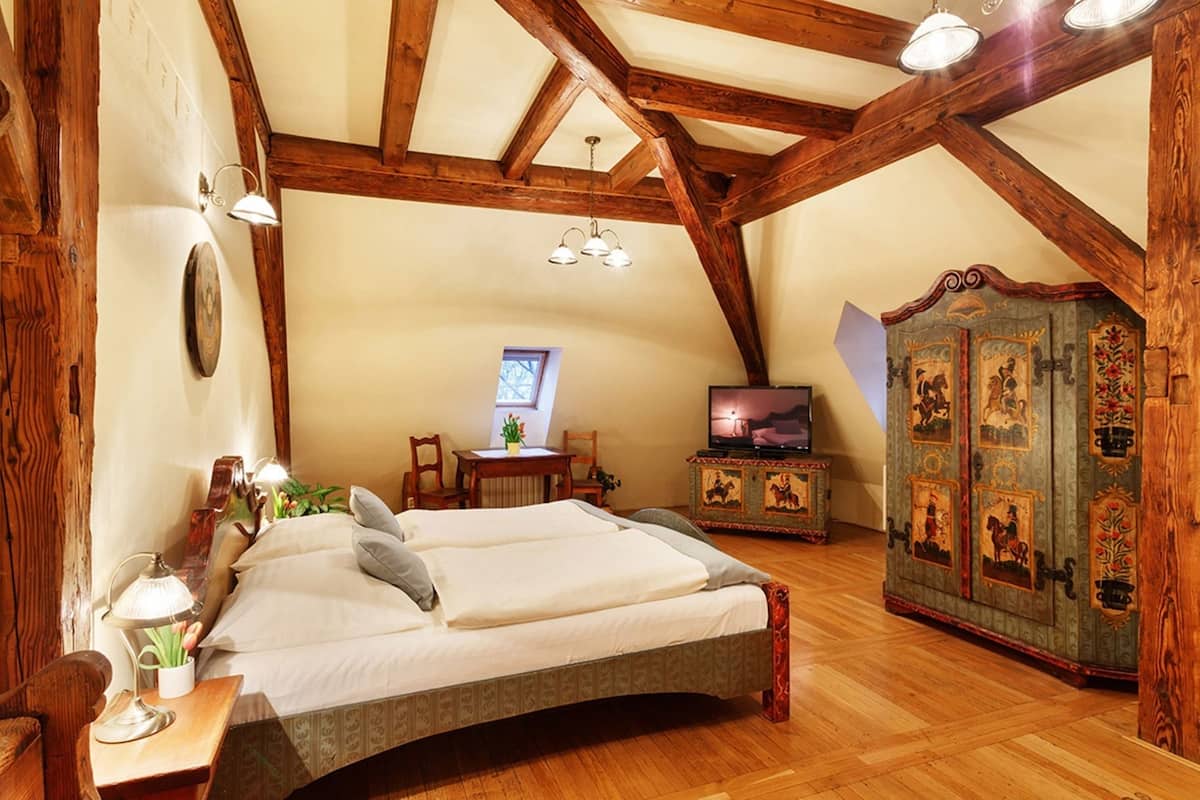 A bedroom with a bed, wooden beams, a television, a cabinet with artwork, and a small table with a lamp.