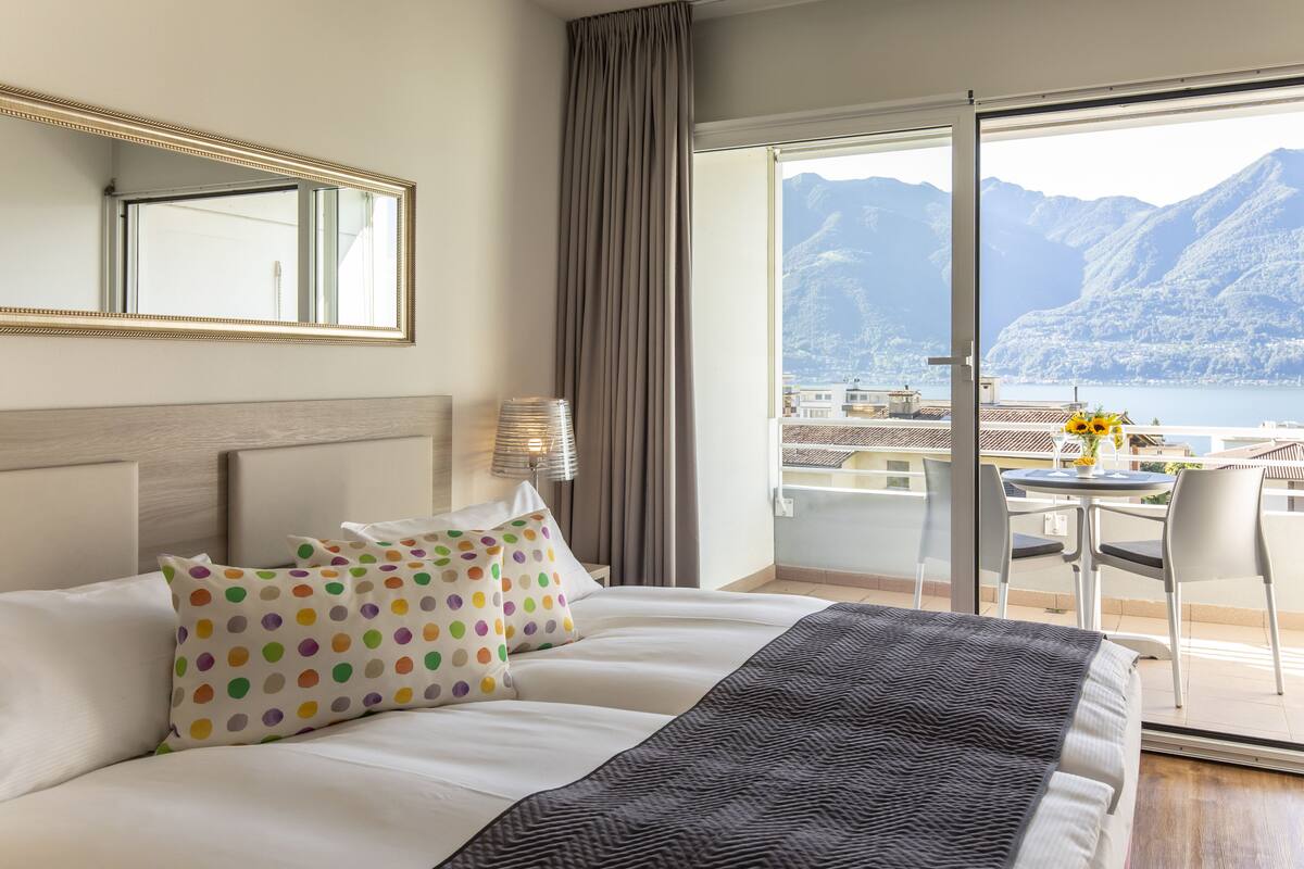 Une chambre d’hôtel comprenant un lit, une table de chevet, une lampe, une chaise et un balcon avec vue sur les montagnes et un lac.