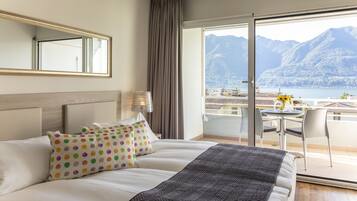 A hotel room with a bed, a nightstand, a lamp, a chair, and a balcony with a view of mountains and a lake.
