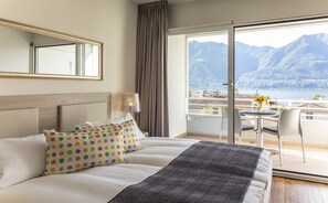A hotel room with a bed, a nightstand, a lamp, a chair, and a balcony with a view of mountains and a lake. - Smart-HOTEL MINUSIO, a Swiss Quality Hotel (Minusio)