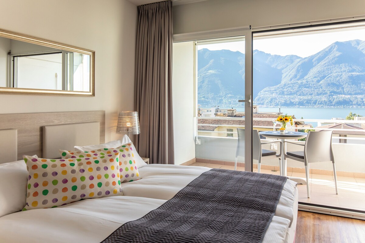 A hotel room with a bed, a nightstand, a lamp, a chair, and a balcony with a view of mountains and a lake.
