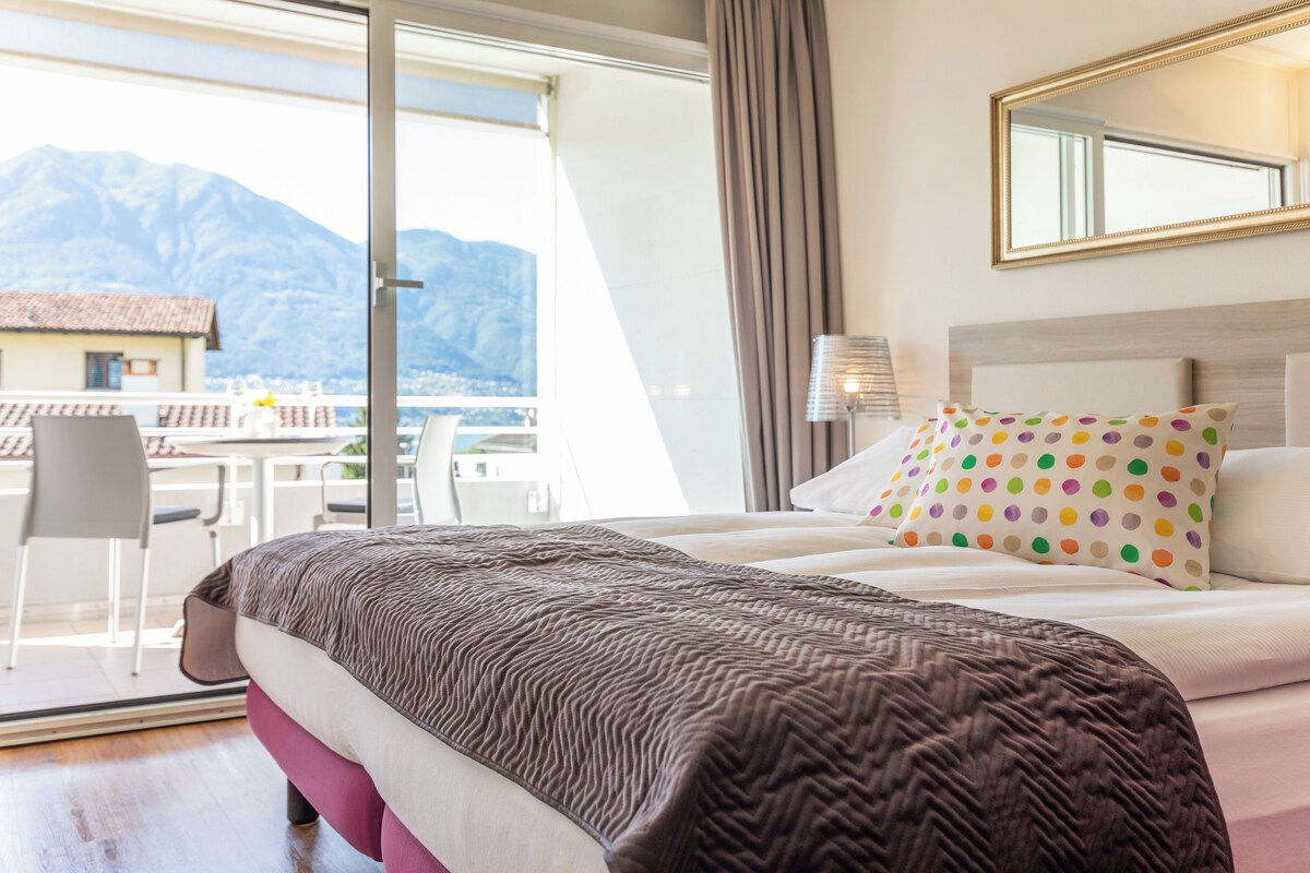 A hotel room with a bed, a window with a view of mountains, a chair, and a table with a view.