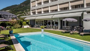 A swimming pool with lounge chairs and umbrellas, surrounded by a well-maintained lawn and a building with large windows and balconies. - Smart-HOTEL MINUSIO, a Swiss Quality Hotel (Minusio)