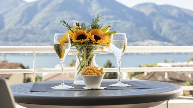 A table with a vase of sunflowers and two glasses of wine, a bowl of snacks, and a view of mountains and water. - Smart-HOTEL MINUSIO, a Swiss Quality Hotel (Minusio)